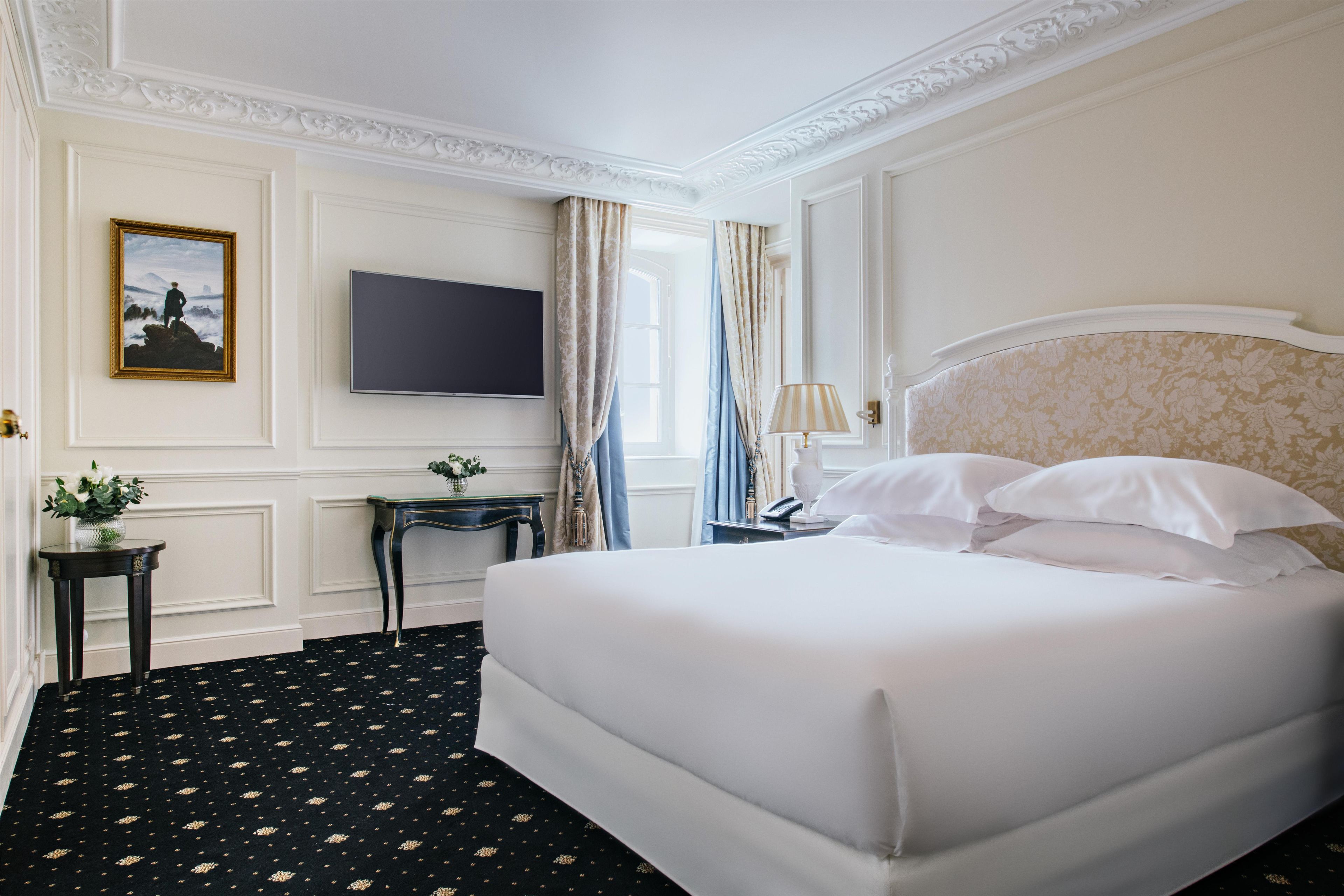 Habitación de hotel con una cama grande, televisor de pantalla plana, una mesita con un florero y una ventana con cortinas.