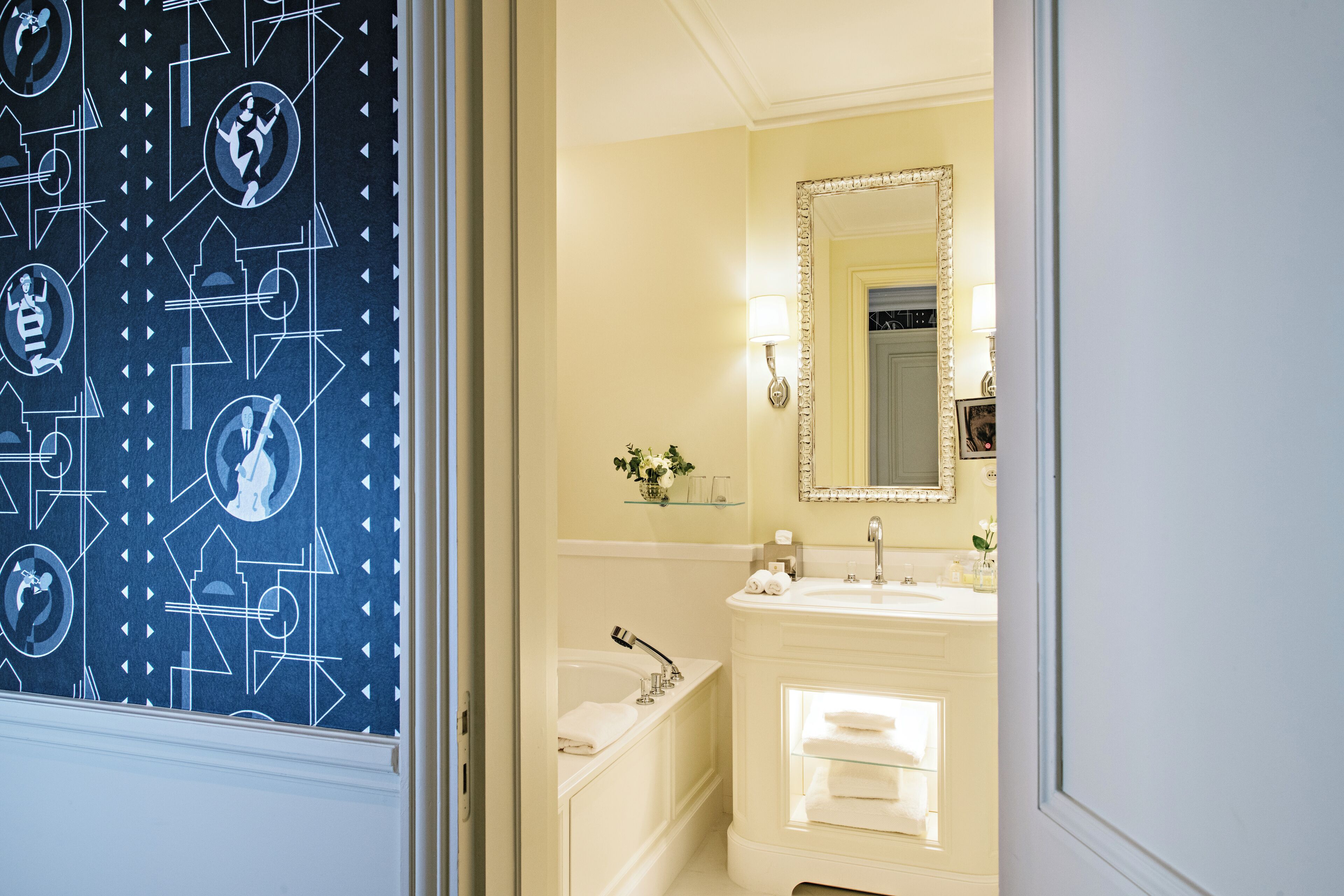 Une salle de bain avec un lavabo blanc, un miroir et une baignoire. On y trouve une étagère avec des articles de toilette et un vase avec des fleurs.
