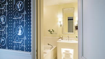 Une salle de bain avec un lavabo blanc, un miroir et une baignoire. On y trouve une étagère avec des articles de toilette et un vase avec des fleurs.