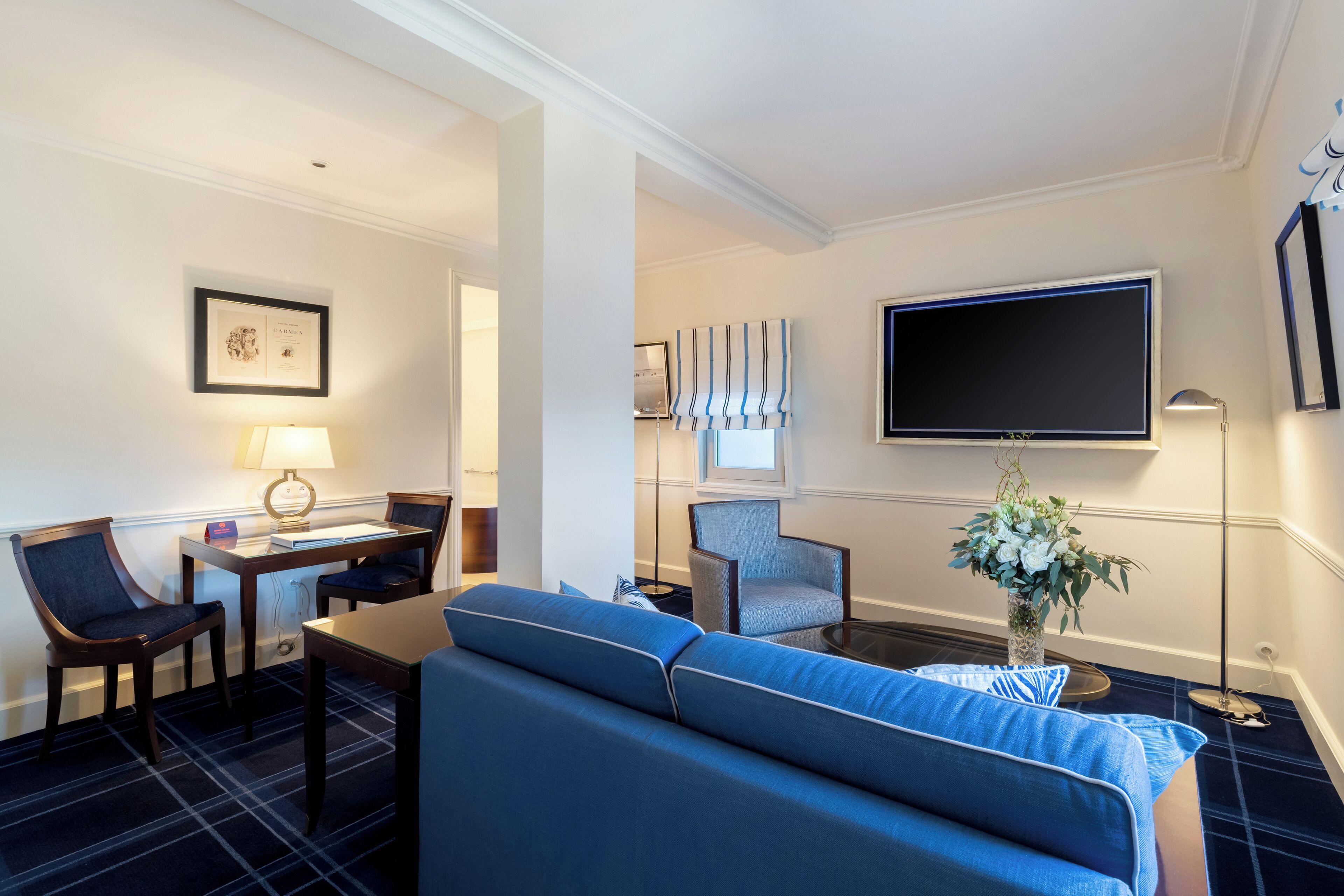 A hotel room with a sofa, chairs, a desk, a flat-screen TV, and a lamp.
