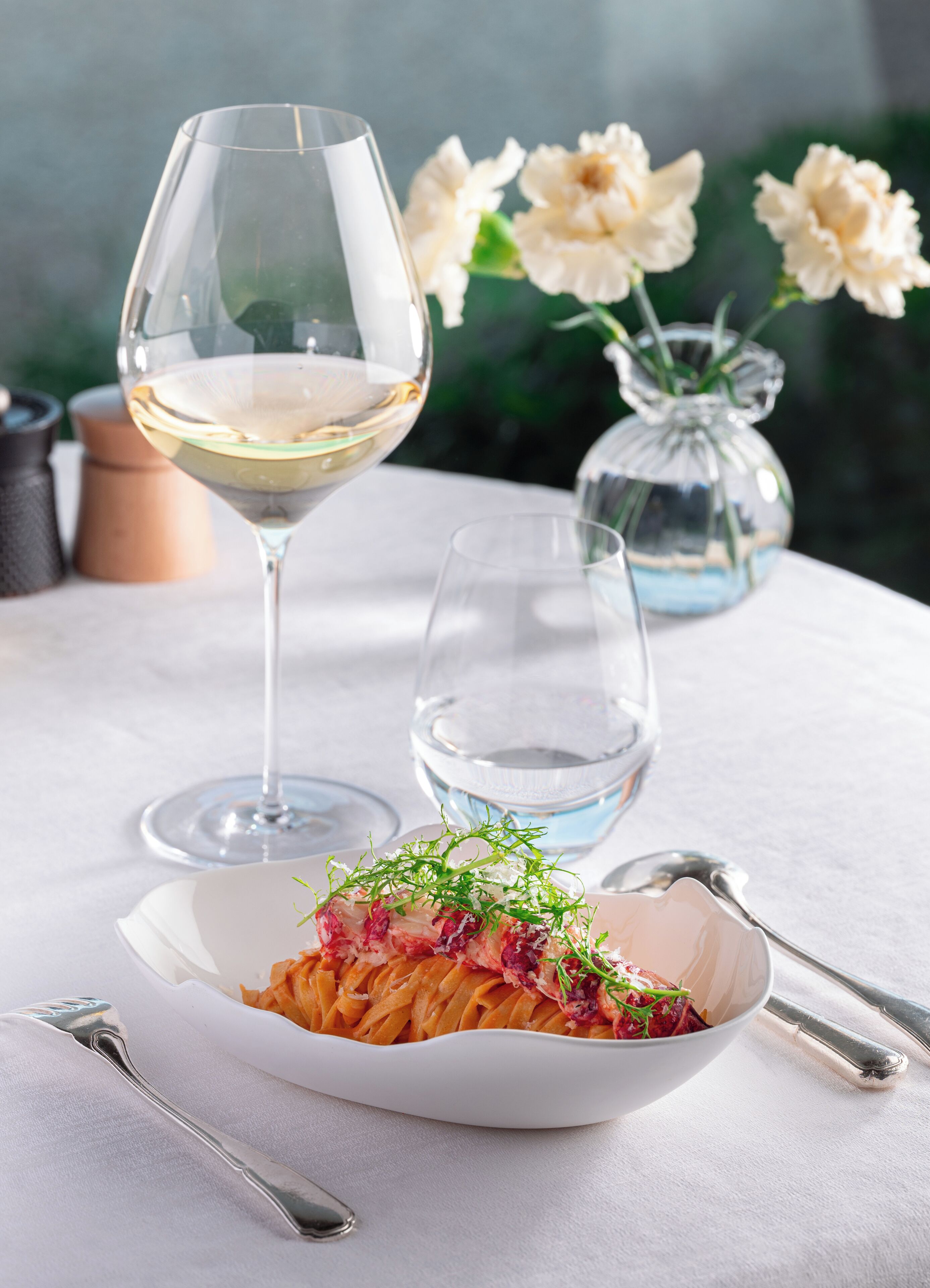 A dining table with a white plate of pasta garnished with greens, a glass of white wine, and a clear glass of water.
