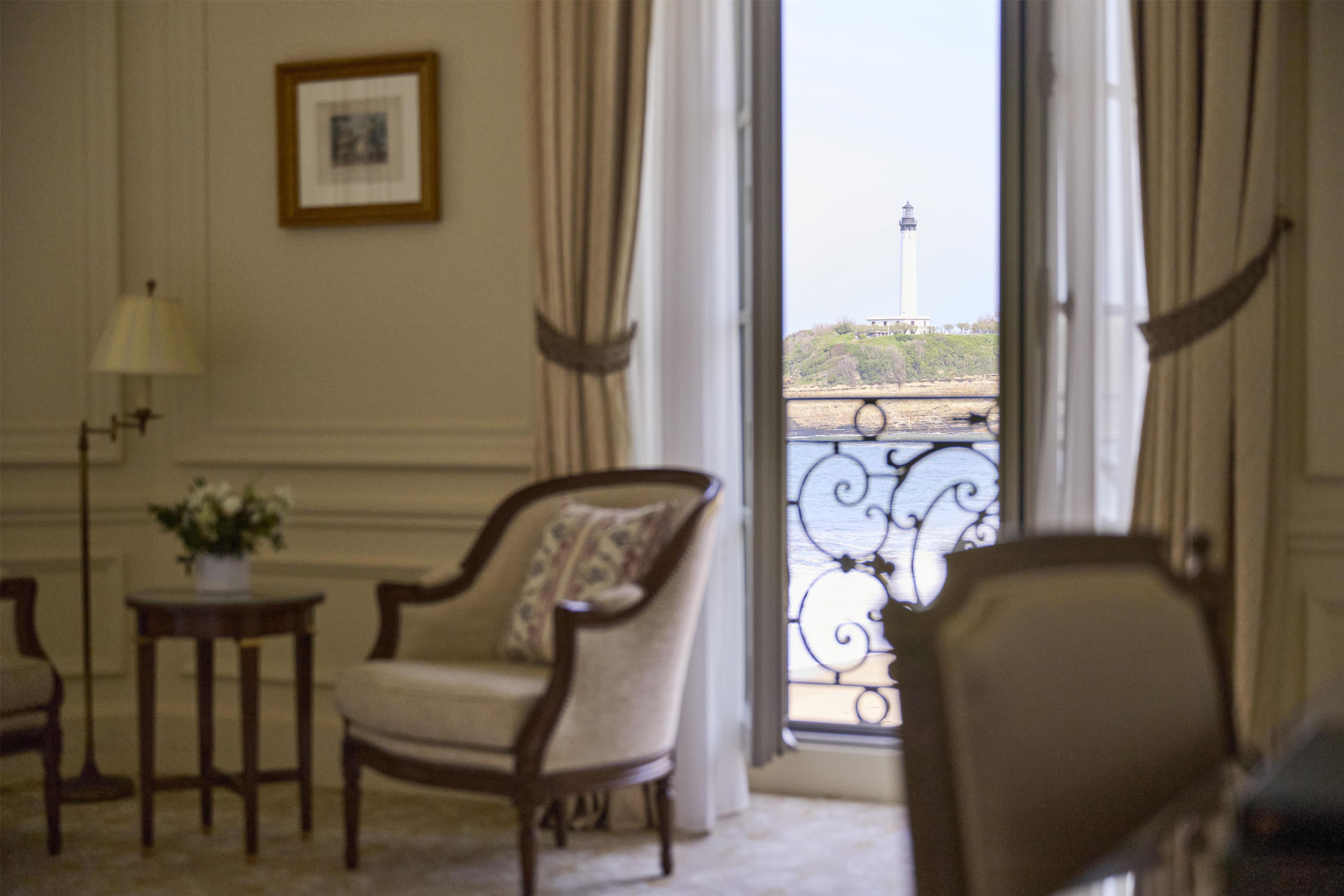 Habitación con vista a un faro a través de una ventana. Se observa una silla, una mesita con una lámpara y una planta en maceta.