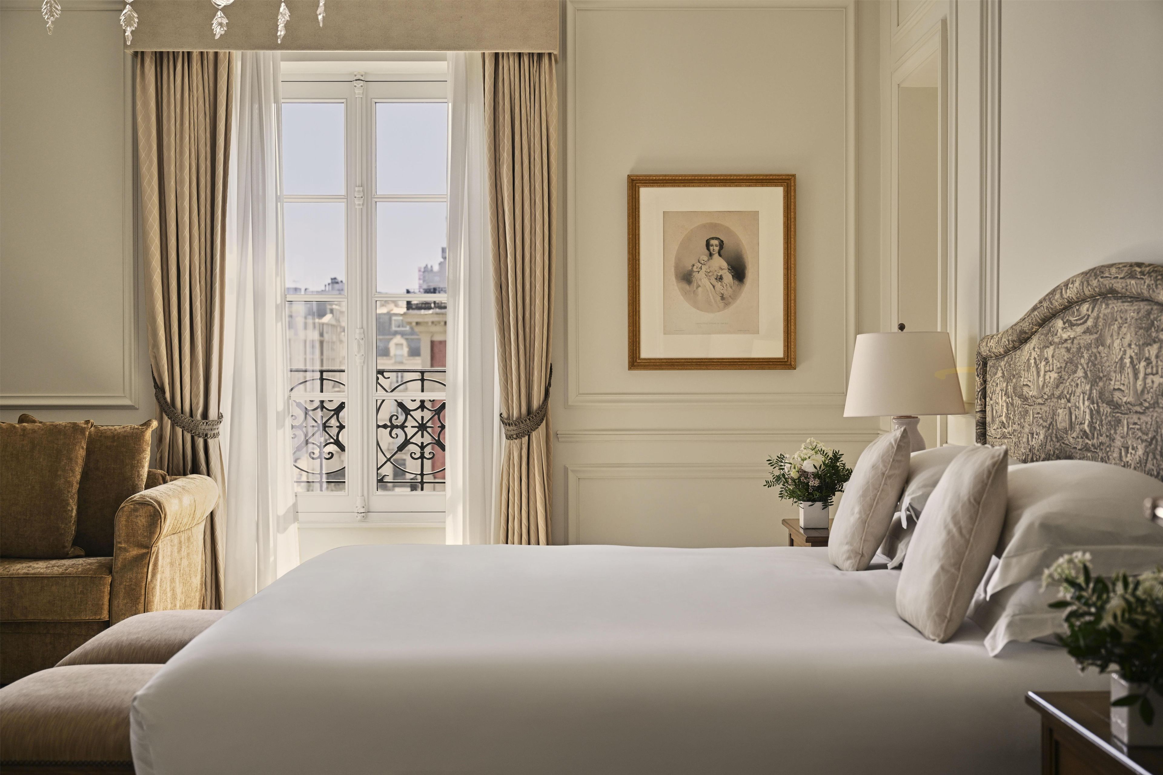 Habitación de hotel con una cama grande, una mesita de noche con lámpara, un retrato enmarcado en la pared y vistas de la ciudad a través de la ventana.