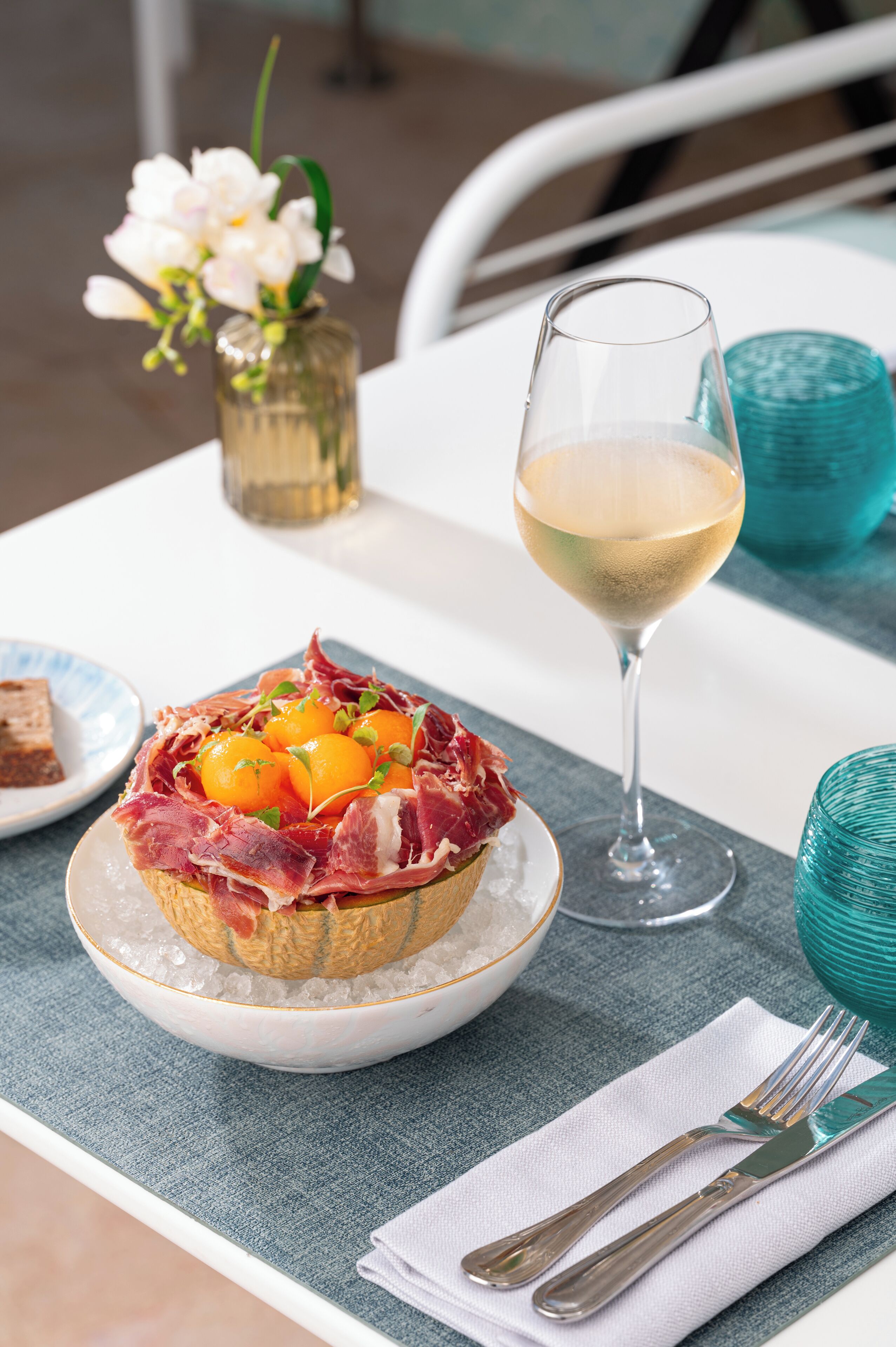 A table setting with a glass of white wine, a plate with a meat dish, and a small vase with flowers.