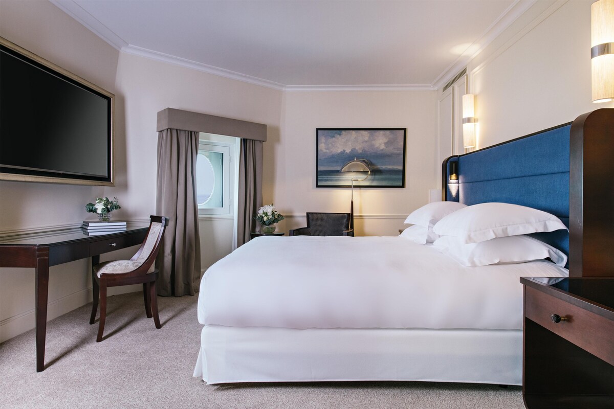 A hotel room with a bed, desk, chair, TV, and a painting on the wall.