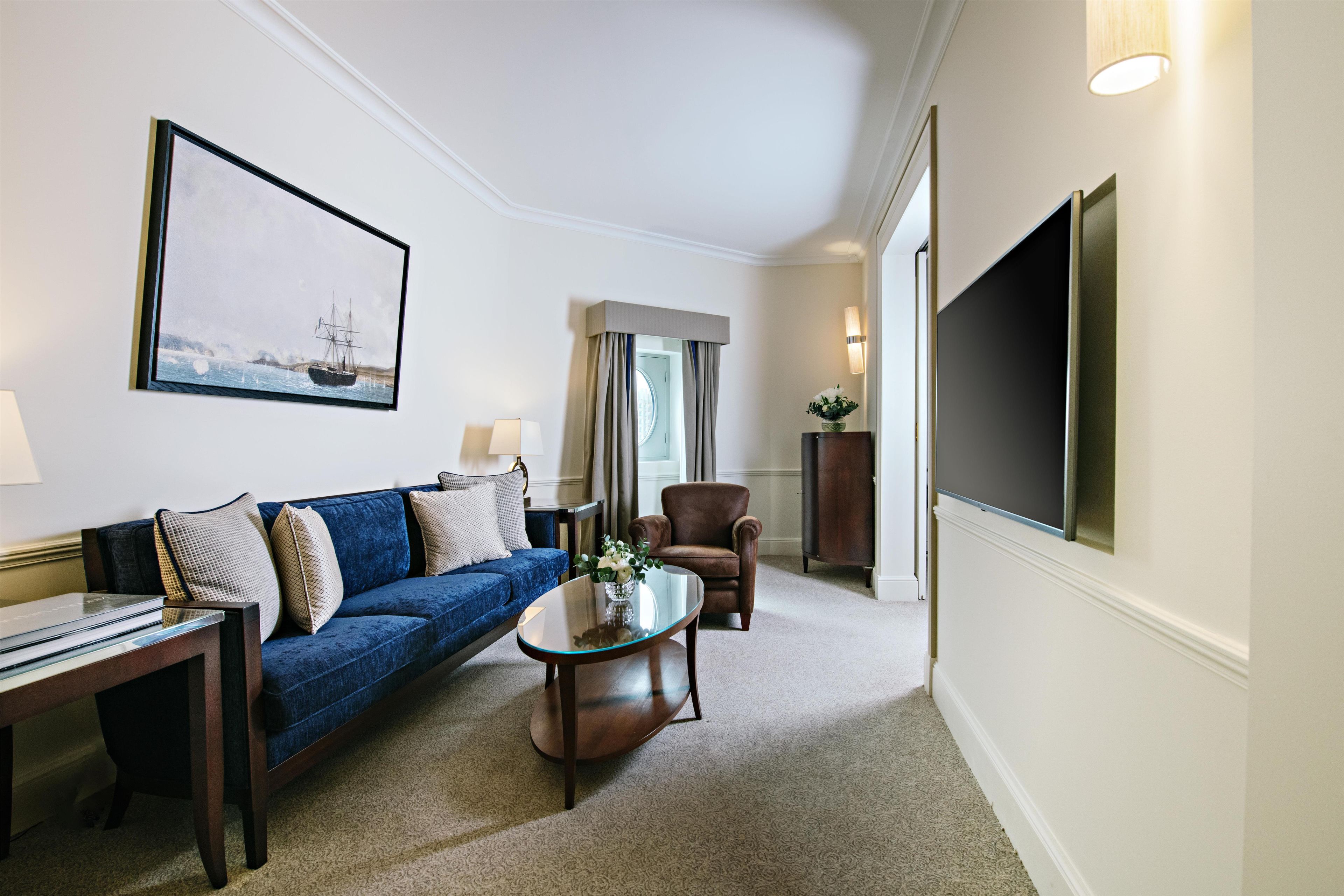A hotel room with a blue sofa, a glass coffee table, a brown armchair, a flat-screen TV, and a painting of a ship on the wall.