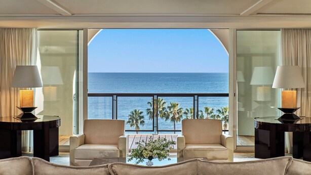 Una habitación lujosa con un ventanal grande que ofrece vistas al mar y palmeras. Hay dos sillones y una mesita con un florero.
