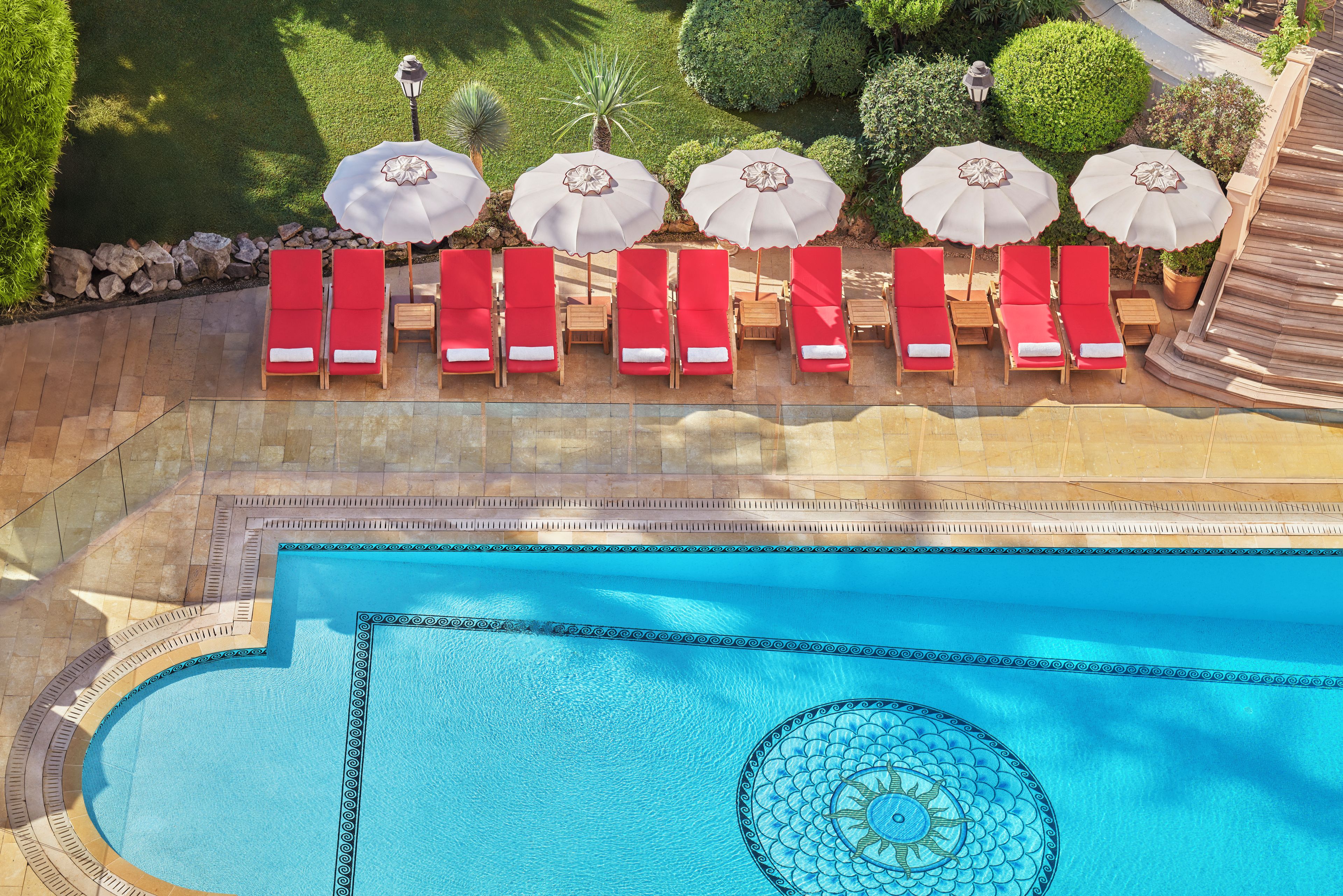 Un área de piscina con sillas reclinables rojas y sombrillas blancas, rodeada por un solárium de piedra y vegetación.