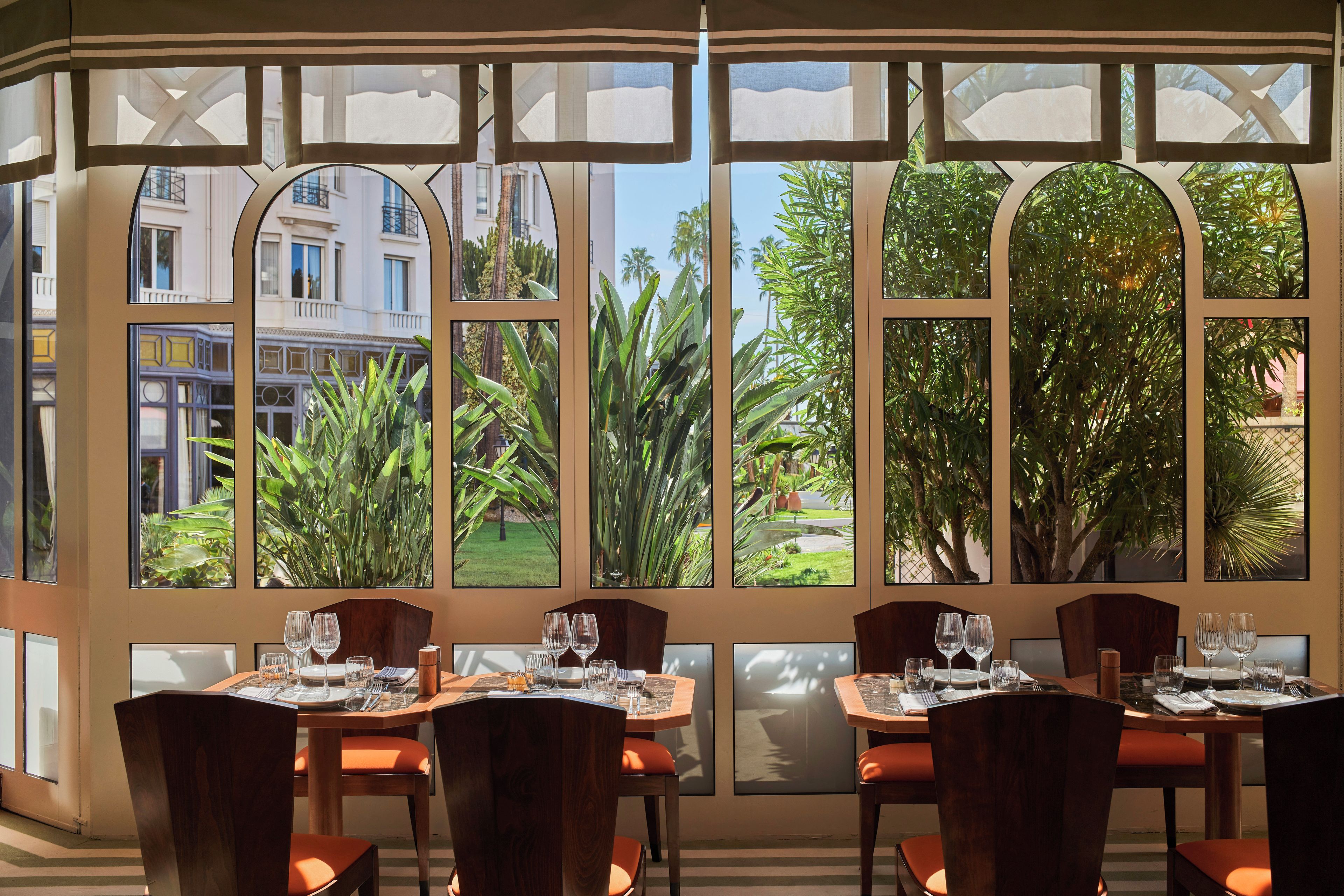 A restaurant with large windows offering views of buildings and greenery. Tables are set with wine glasses and plates.