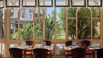 A restaurant with large windows offering views of buildings and greenery. Tables are set with wine glasses and plates.