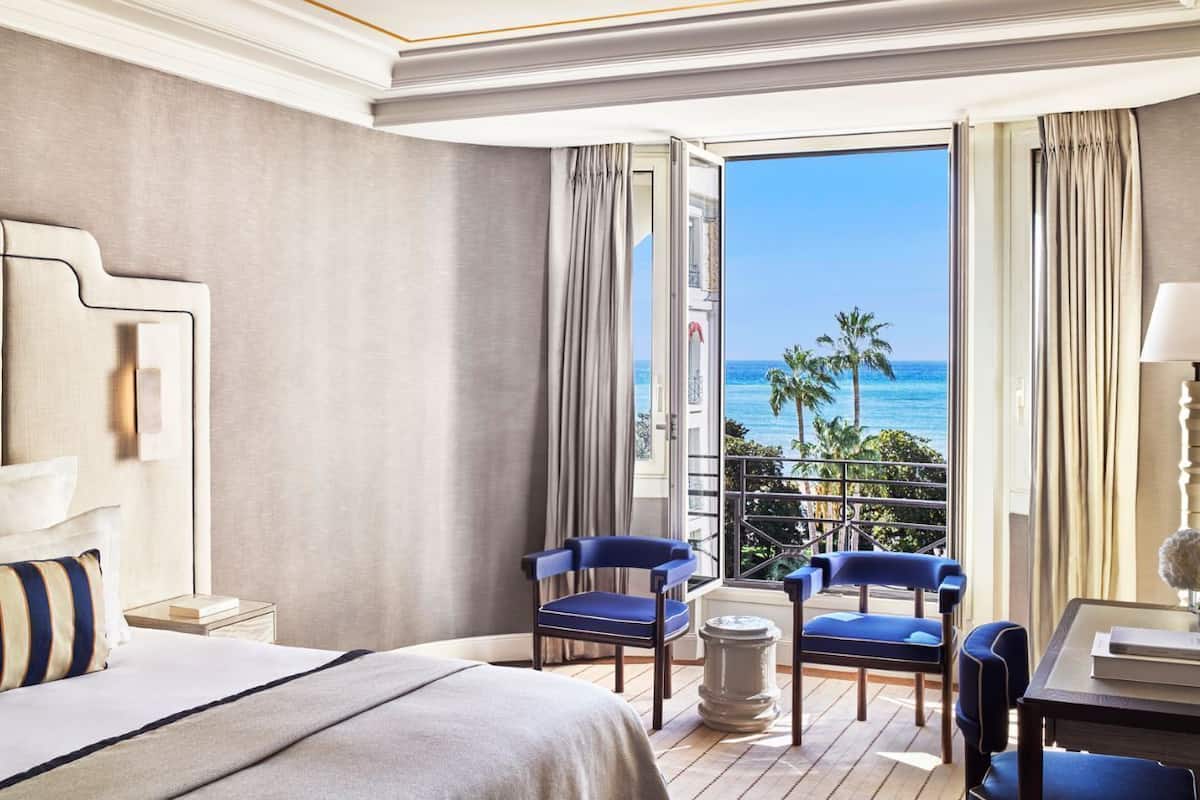 Habitación de hotel con una cama, dos sillas azules, una mesita y vista al océano.