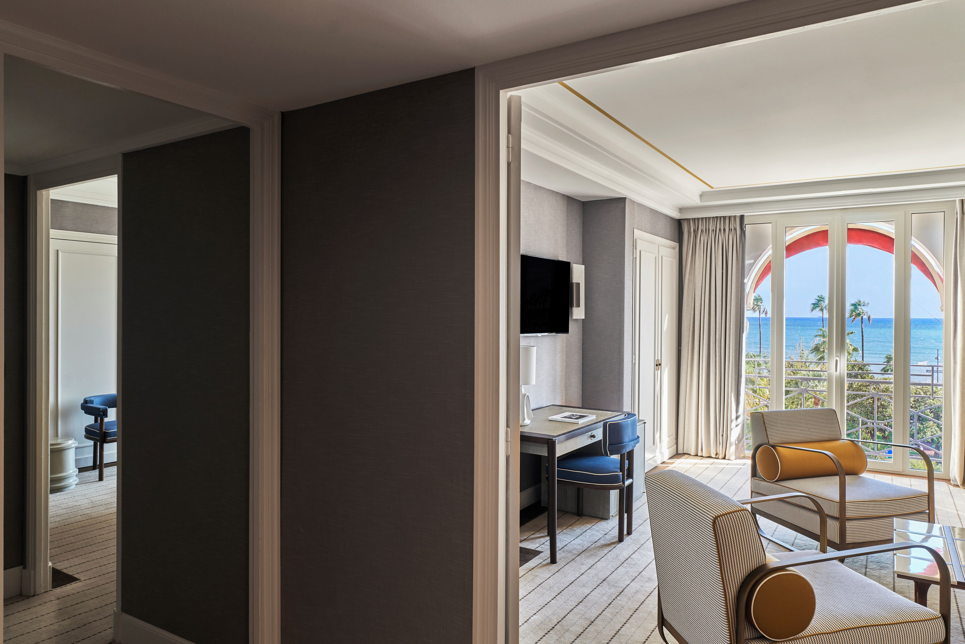 A hotel room with a balcony, a desk, and a seating area with cushions.