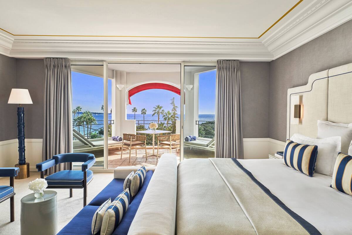 Una habitación de hotel con una cama grande, una zona de estar con vista al océano y un balcón con mesa y sillas.