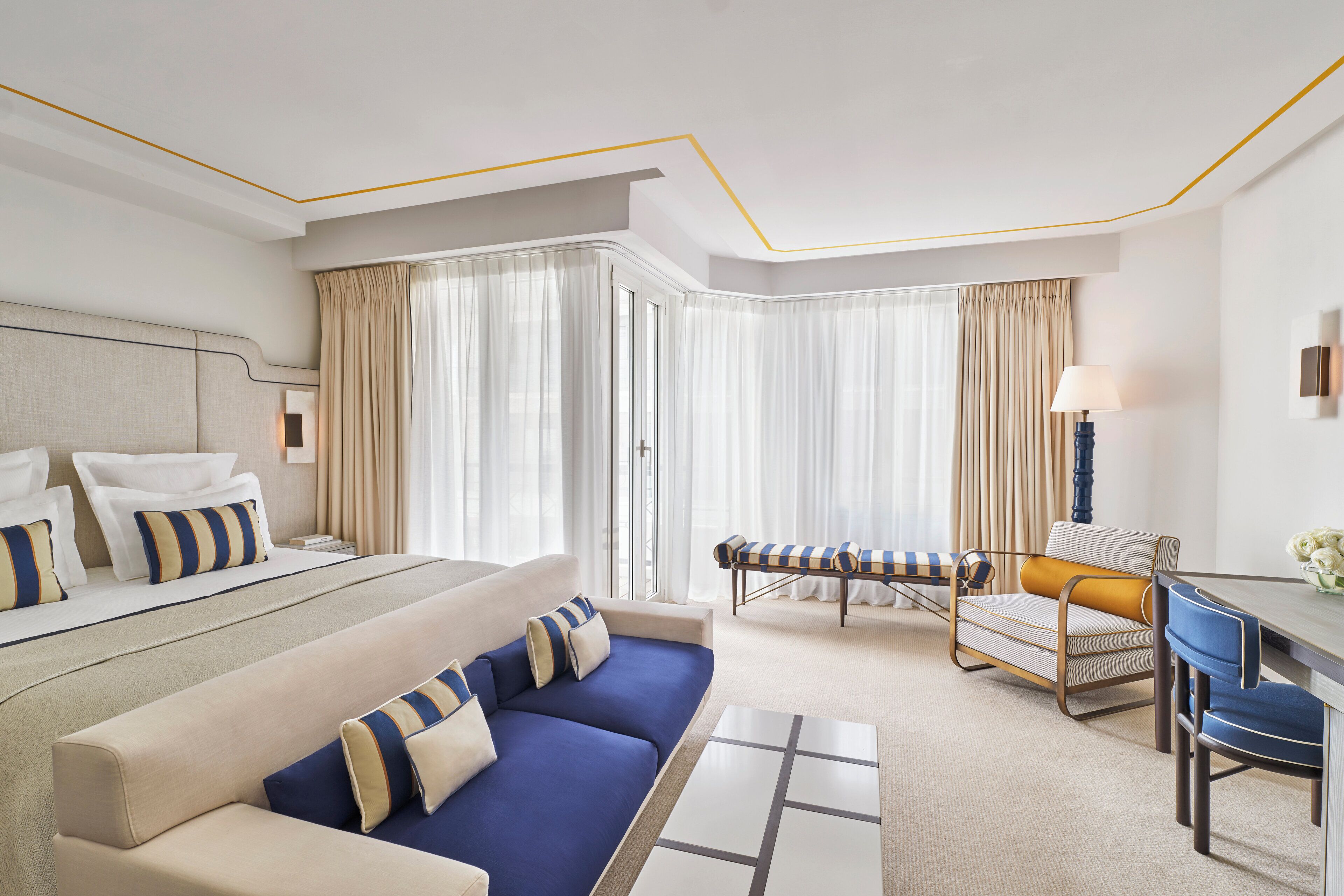 A modern hotel room with a large bed, a blue sofa, a small table, and a desk with a lamp.