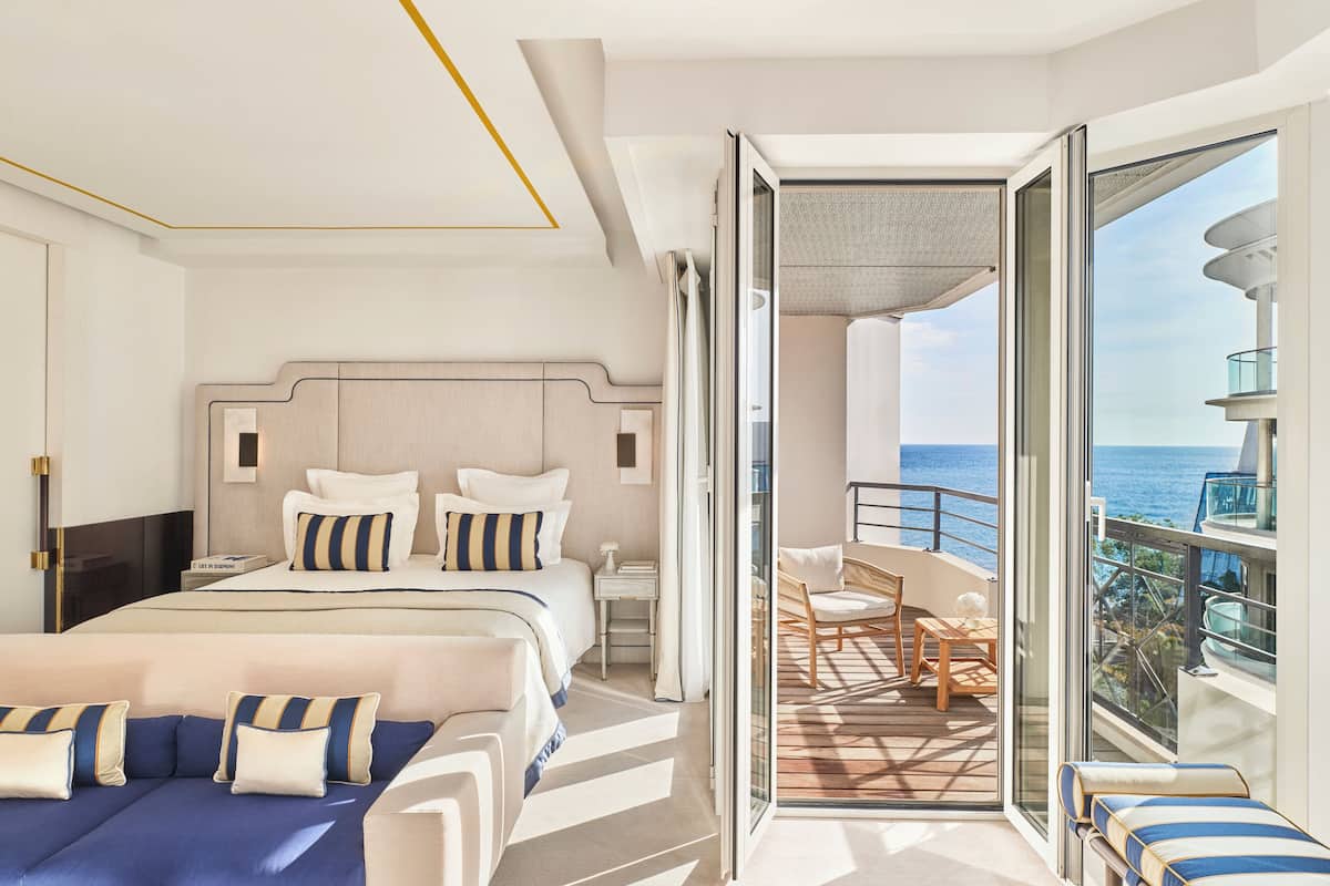 Una habitación de hotel moderna con una cama grande, un balcón con vistas al océano y una zona de estar separada con una mesa y sillas.