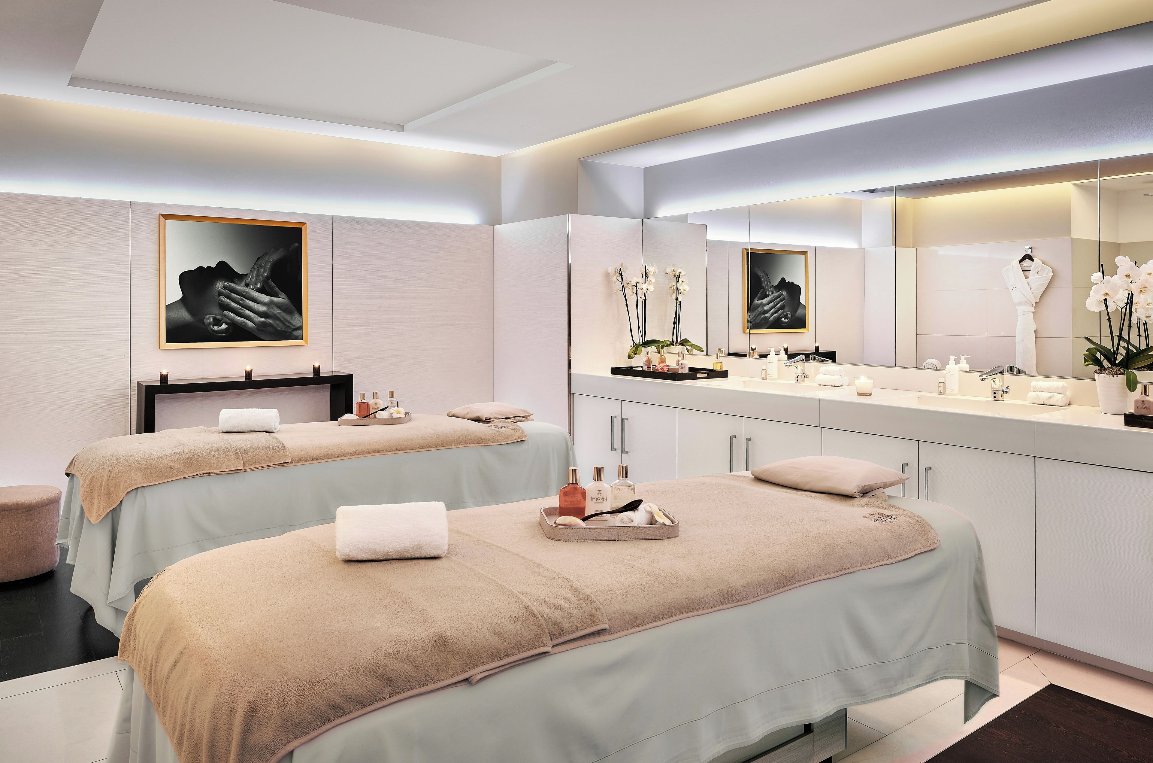 A modern spa room with two massage tables, a large mirror, and a vanity with toiletries.