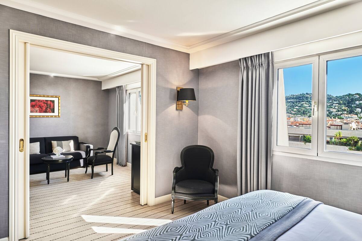 Una habitación de hotel moderna con una cama, una silla, un sofá, una mesita y vistas a la ciudad a través de amplias ventanas.