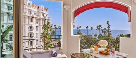 A balcony with a view of the sea, a dining table with breakfast, and a lounge area with a cushion.