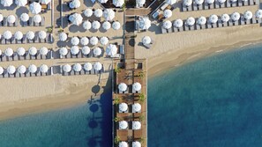 Vue aérienne d’un complexe hôtelier en bord de mer, avec plusieurs rangées de chaises longues et de parasols, une terrasse en bois et une vue dégagée sur la plage et l’océan adjacents.