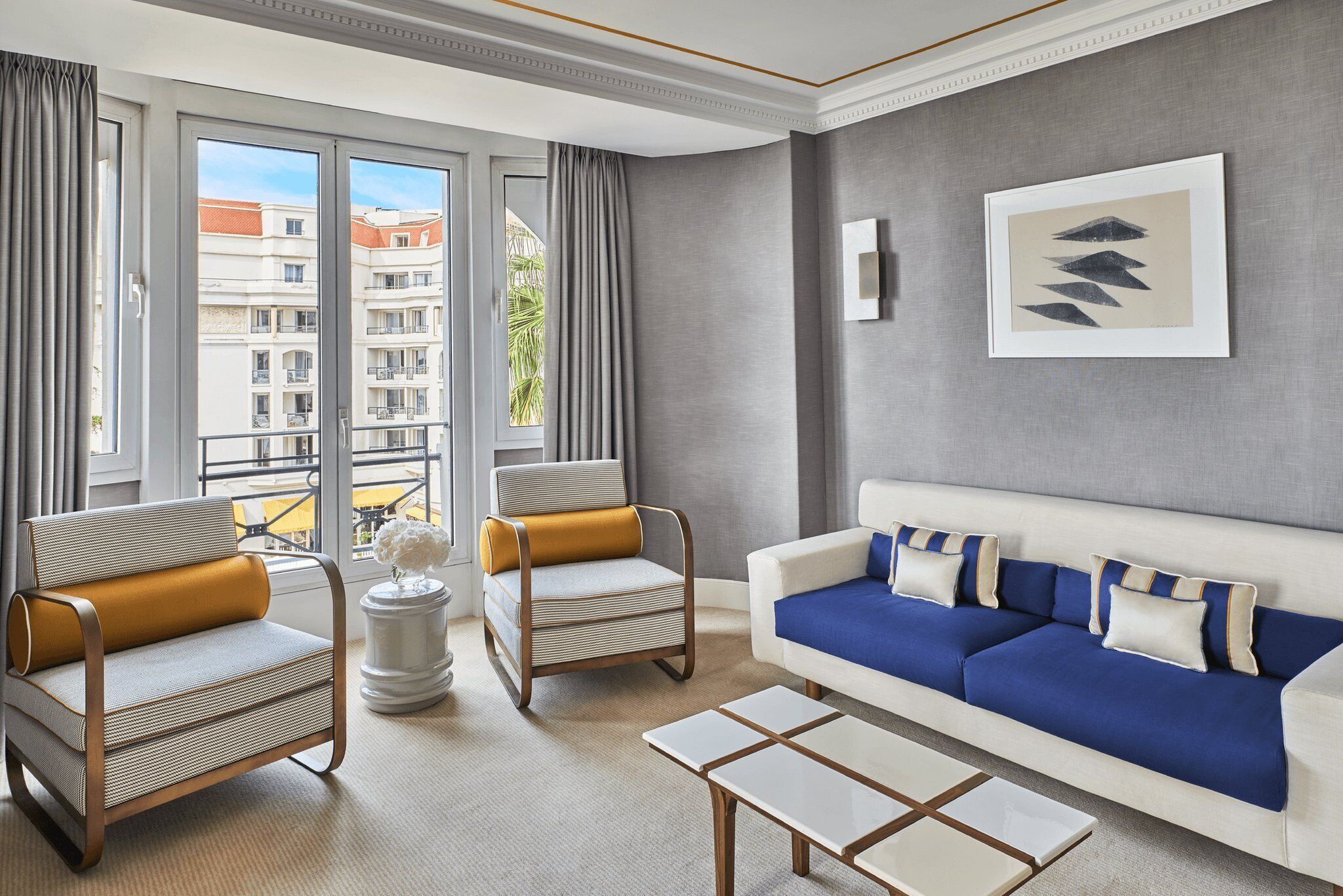 Un salon moderne comprenant un canapé, deux fauteuils, une table basse et une vue sur un immeuble grâce à de grandes fenêtres.