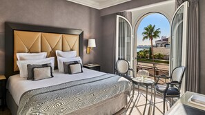 Habitación de hotel con una cama grande, un área de descanso con una mesa y sillas, vista de la ciudad a través de un ventanal amplio y un balcón con una palmera.