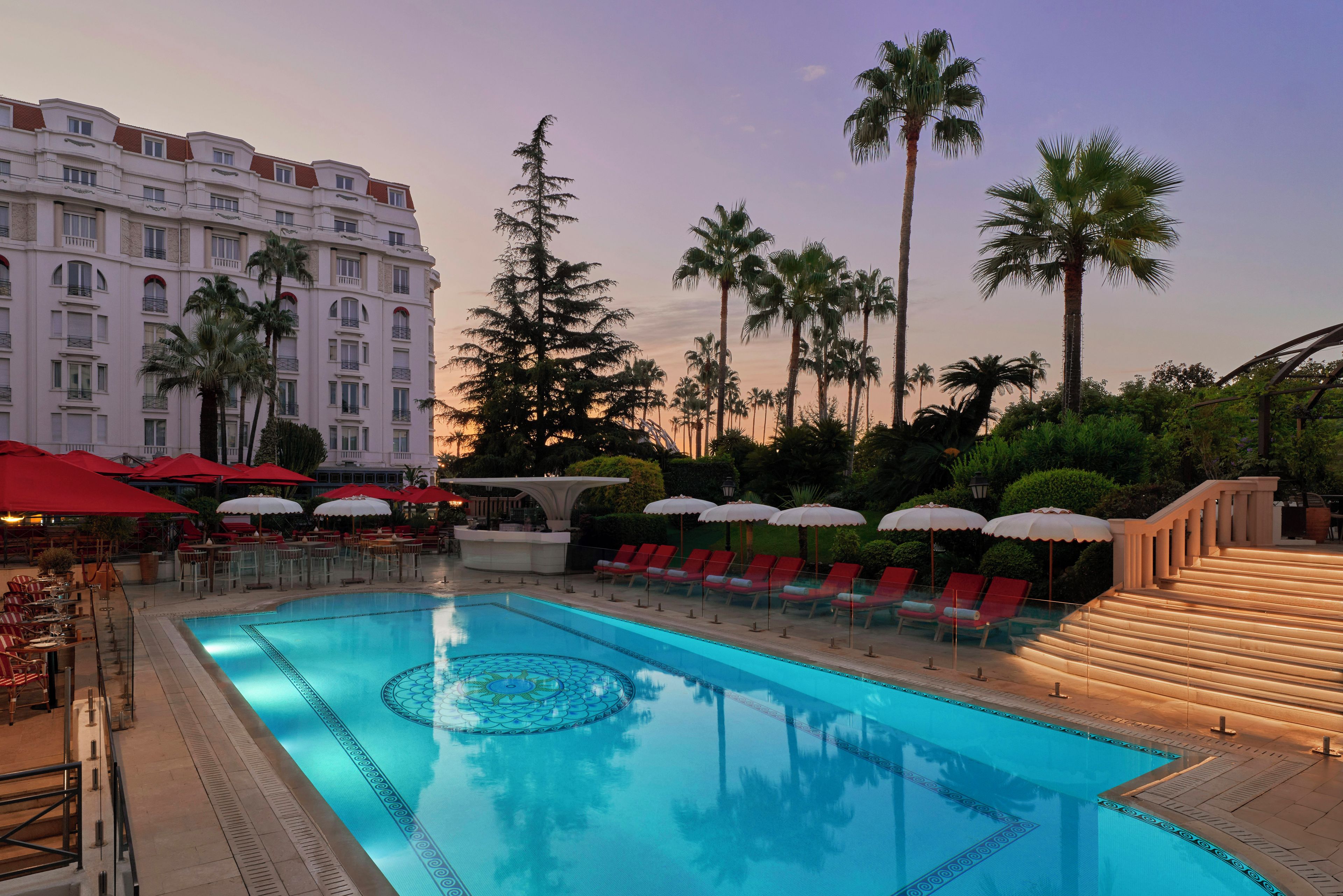 A hotel pool with a geometric pattern, surrounded by palm trees and a building with balconies.