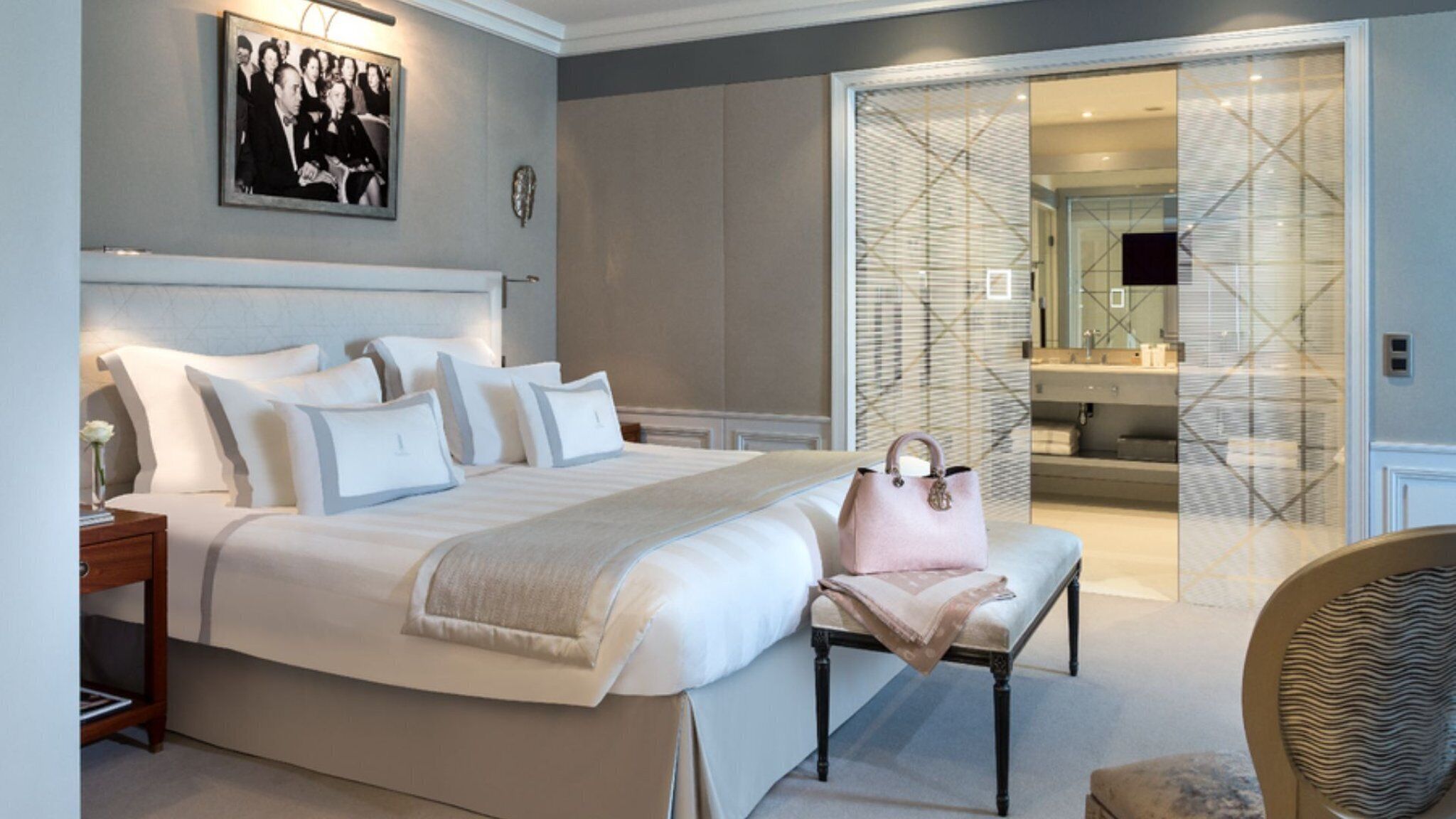 A bedroom with a large bed, bedside tables, a bench, a handbag, and a mirror.