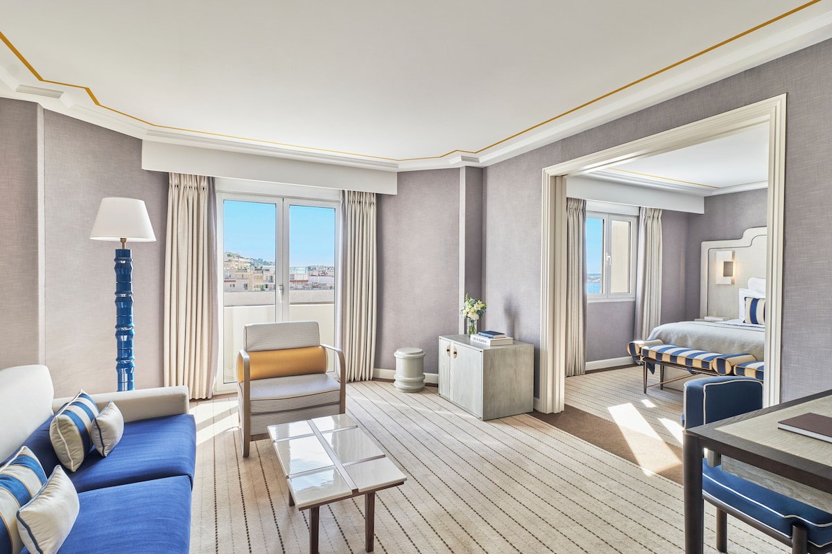 Une chambre d’hôtel avec un canapé, un fauteuil et une table à manger. On y trouve une grande fenêtre avec des rideaux, un petit meuble et un lit avec une tête de lit.