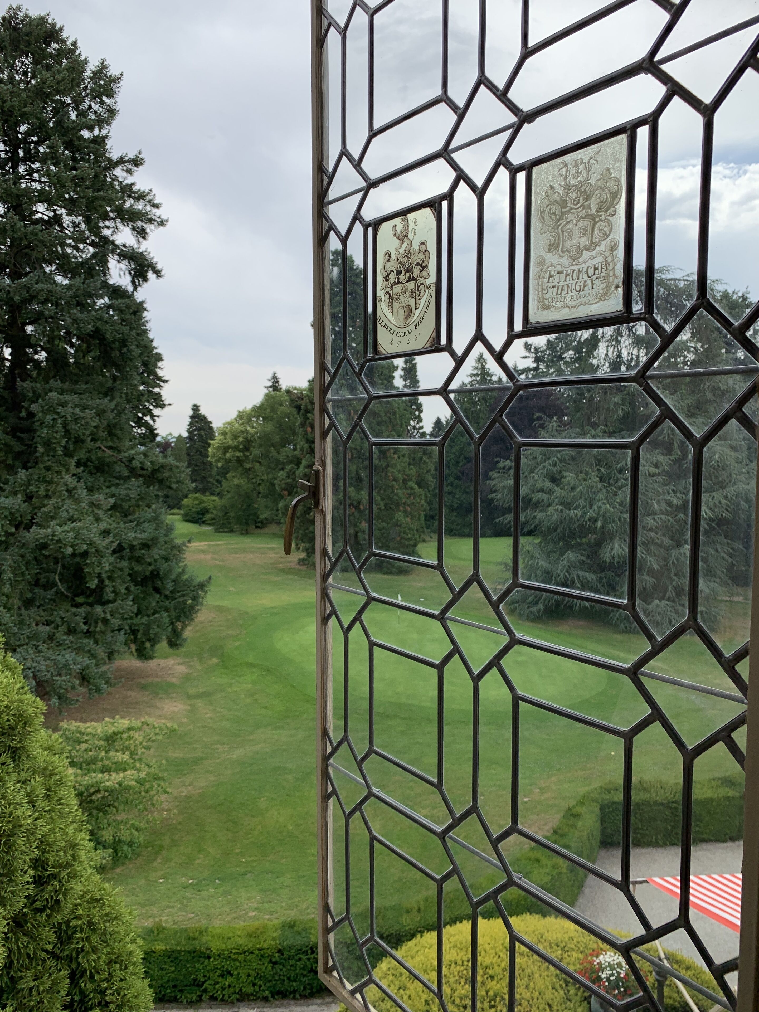 Vista a través de una ventana de diseño geométrico con emblemas heráldicos, un césped cuidado y árboles frondosos.
