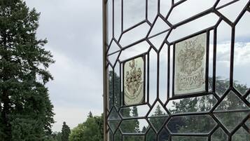 Vista a través de una ventana de diseño geométrico con emblemas heráldicos, un césped cuidado y árboles frondosos.