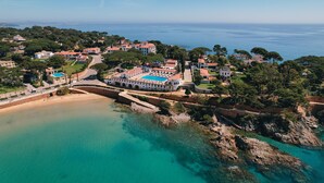 An aerial view of a coastal resort with a large building featuring a swimming pool, surrounded by lush greenery and a clear view of the sea.