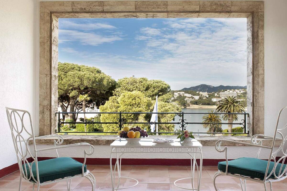 Ein Balkon mit zwei weißen Metallstühlen, einem kleinen Tisch und Blick auf einen See, Bäume und Gebäude in der Ferne.