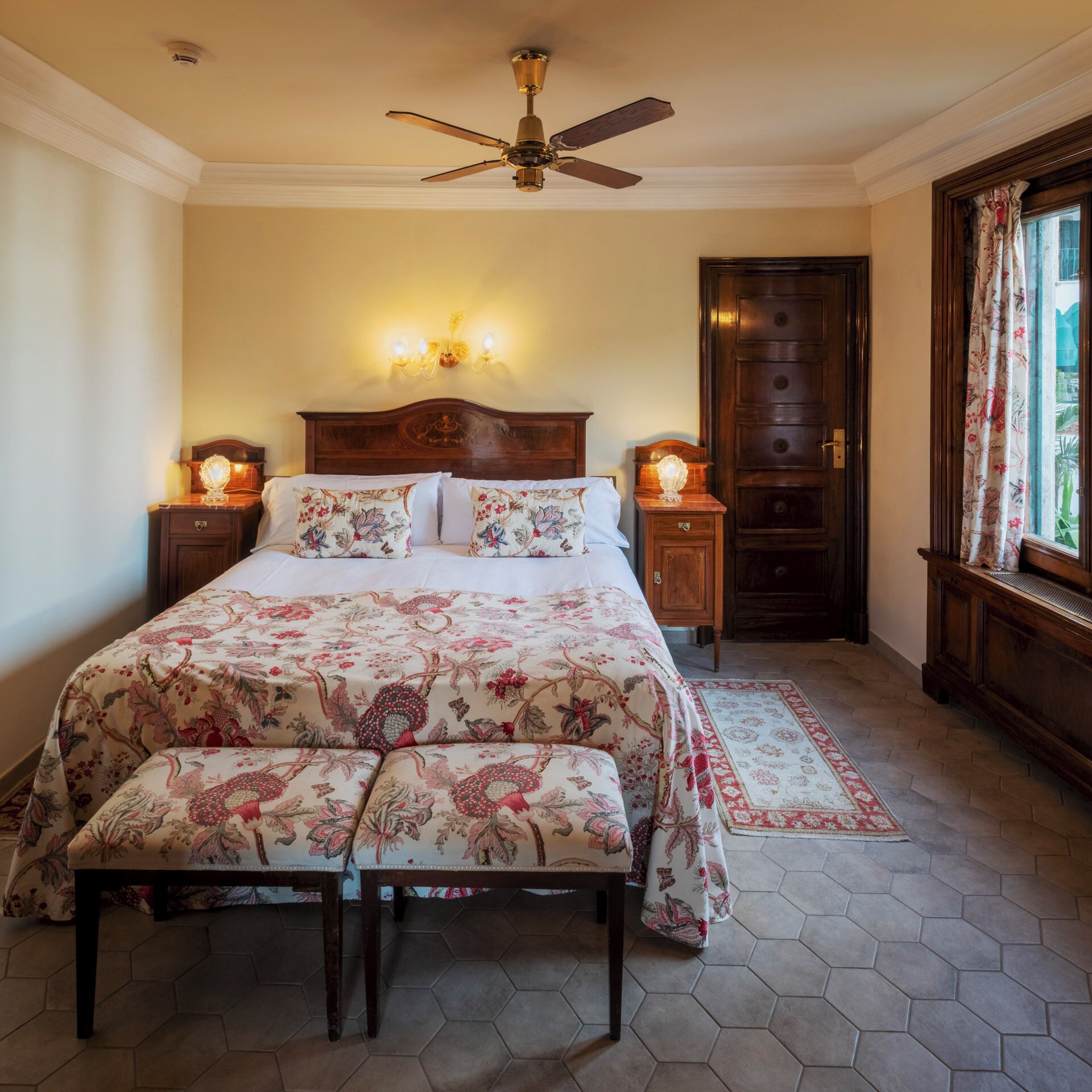 Une chambre avec un grand lit, un ventilateur de plafond, deux tables de chevet avec des lampes, une porte en bois et une fenêtre avec des rideaux.