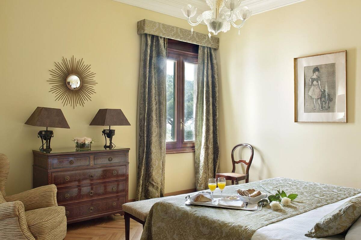 Ein Hotelzimmer mit Bett, Schminktisch, zwei Lampen, einem Kronleuchter, einem Fenster mit Vorhängen, einem gerahmten Bild und einem Frühstücksbrettchen auf dem Bett.