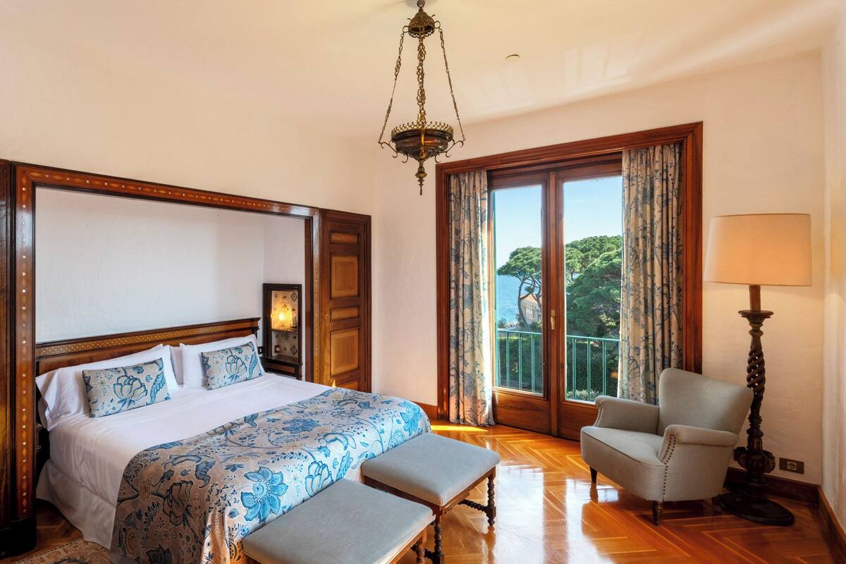 Ein Hotelzimmer mit einem großen Bett, zwei passende Bänke, einem Sessel, einer Lampe und Meerblick vom Balkon.