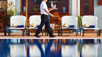 Un hombre caminando cerca de una piscina con sillones de descanso y sombrillas, un bar con botellas, y un cartel que dice "LA".