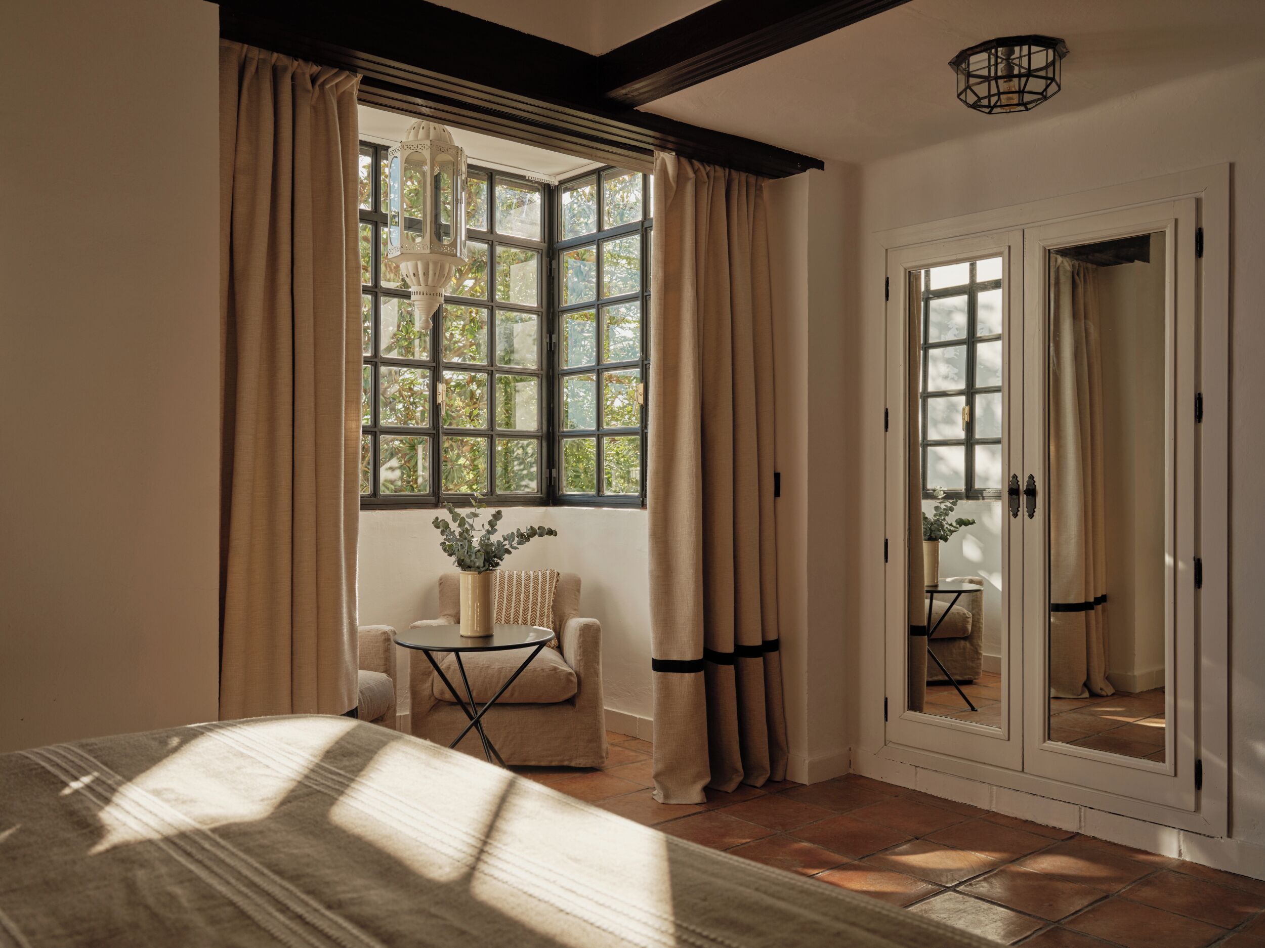 Una habitación con un ventanal grande, una cama, una silla, una mesita y una puerta con un espejo.