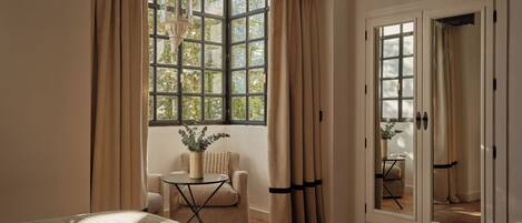 Una habitación con un ventanal grande, una cama, una silla, una mesita y una puerta con un espejo.