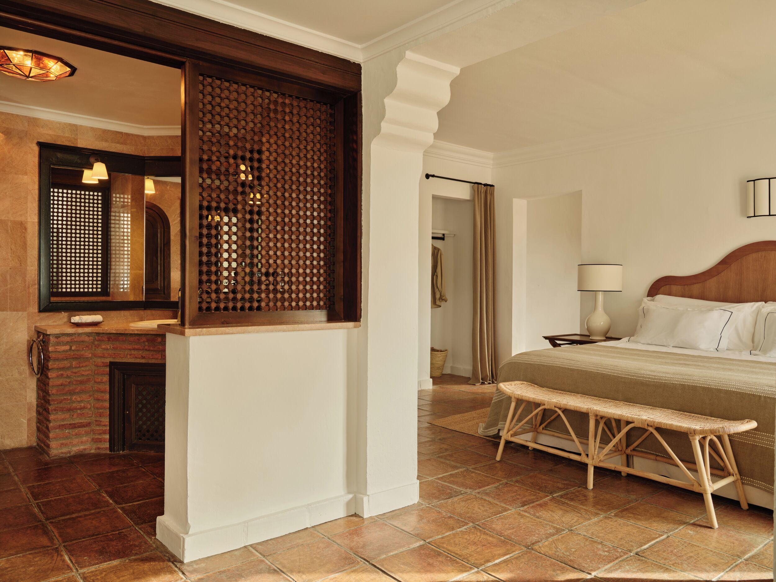 A hotel room with a bed, bedside table, lamp, wicker bench, and a mirror with a wooden frame.