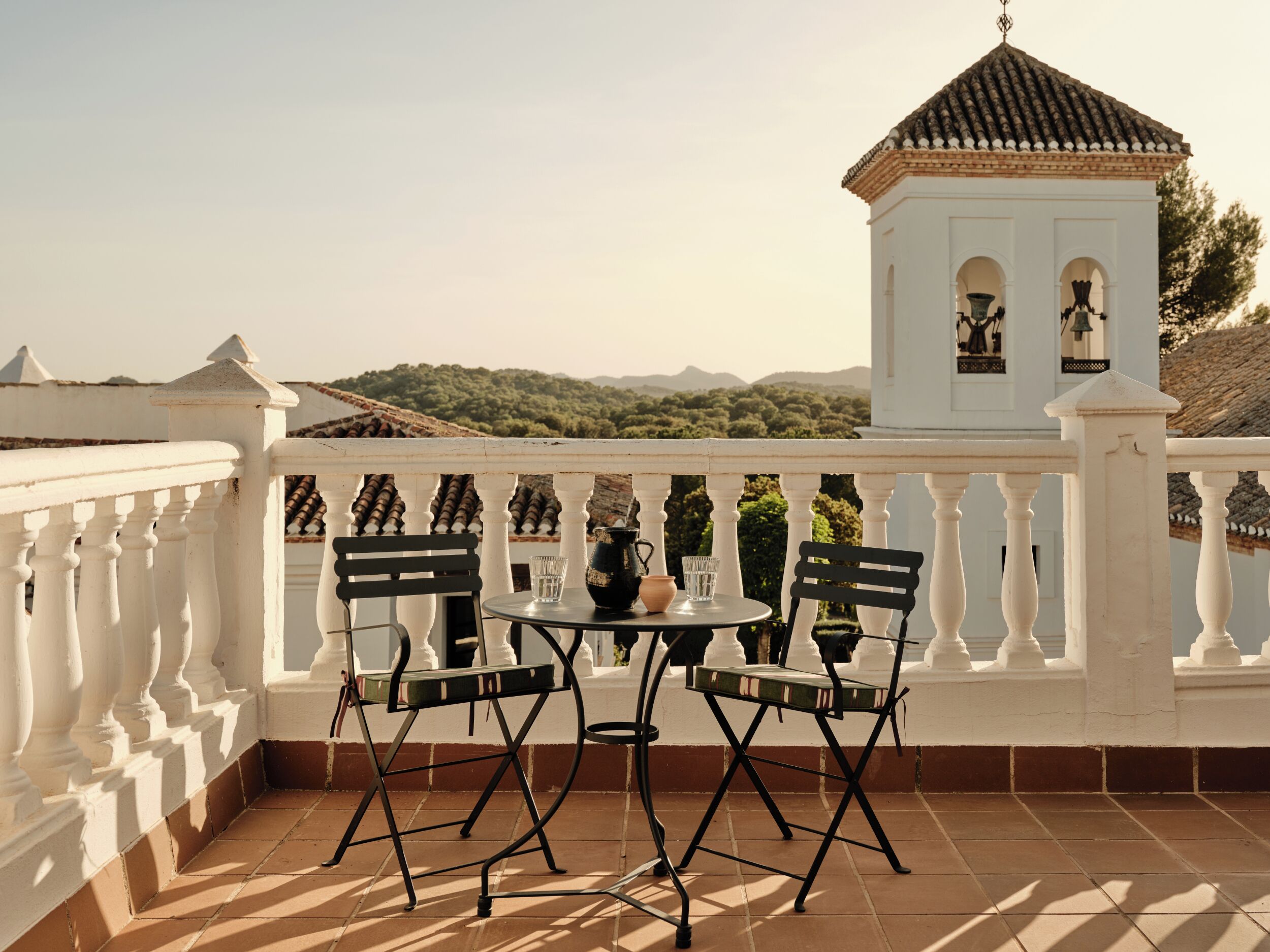 Ein Balkon mit weißer Brüstung, zwei schwarzen Stühlen, einem kleinen runden Tisch und Blick auf ein weißes Gebäude mit Ziegeldach.