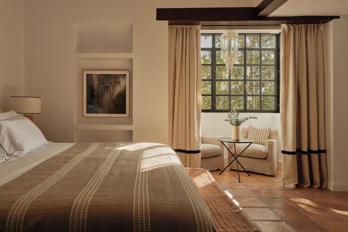 Une chambre à coucher avec un grand lit, une chaise, une petite table, une fenêtre avec des rideaux et un luminaire suspendu.