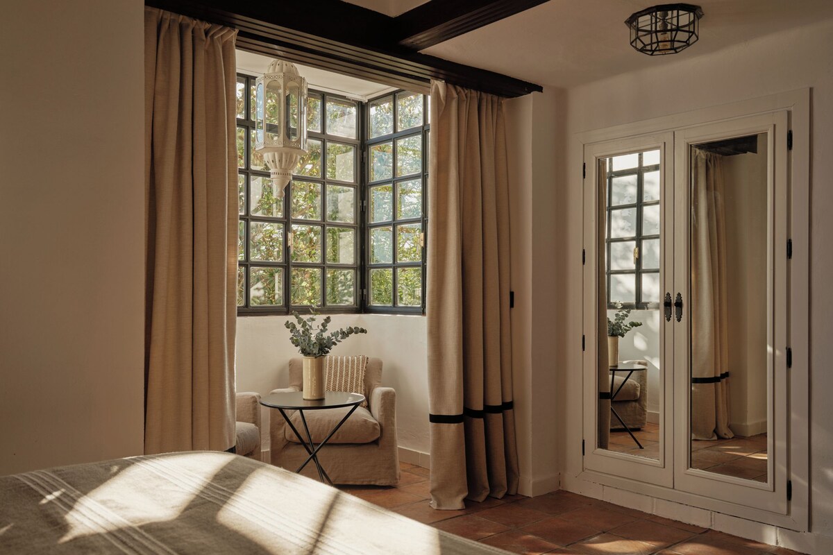 A room with a large window, a bed, a chair, a small table, and a door with a mirror.