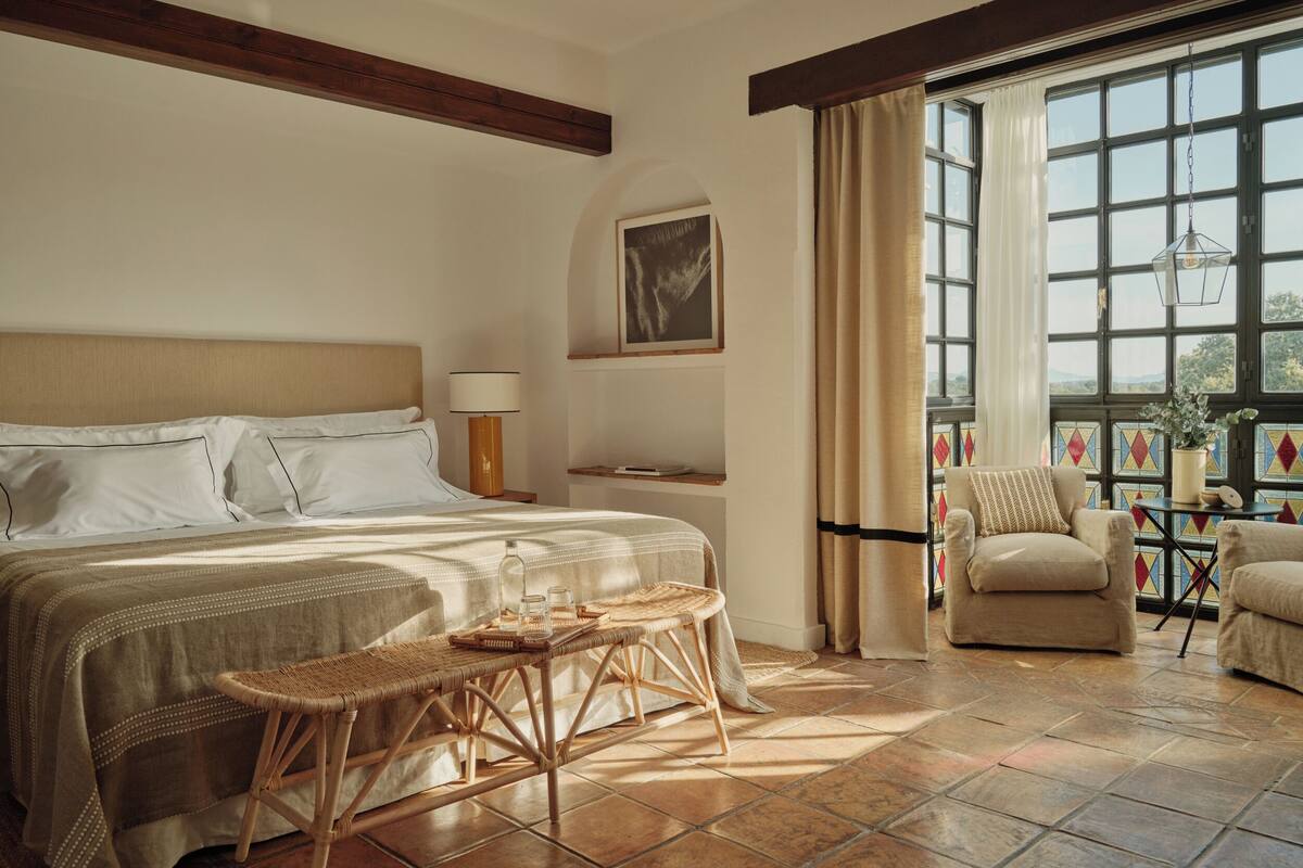 Une chambre à coucher avec un lit, une chaise, une petite table, une lampe et une fenêtre avec des rideaux.