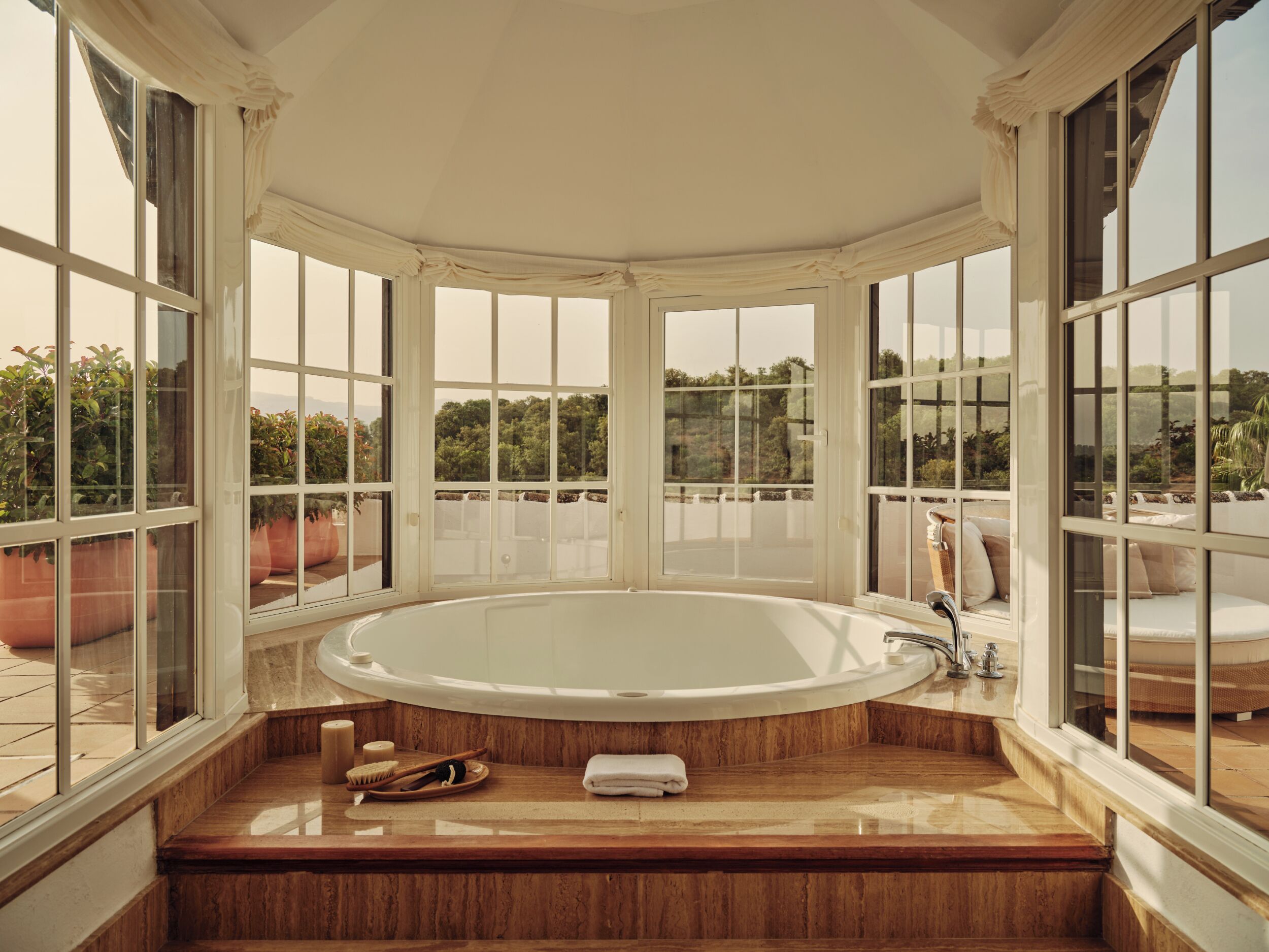 A large, freestanding bathtub in a room with large windows, wooden flooring, and a view of greenery outside.