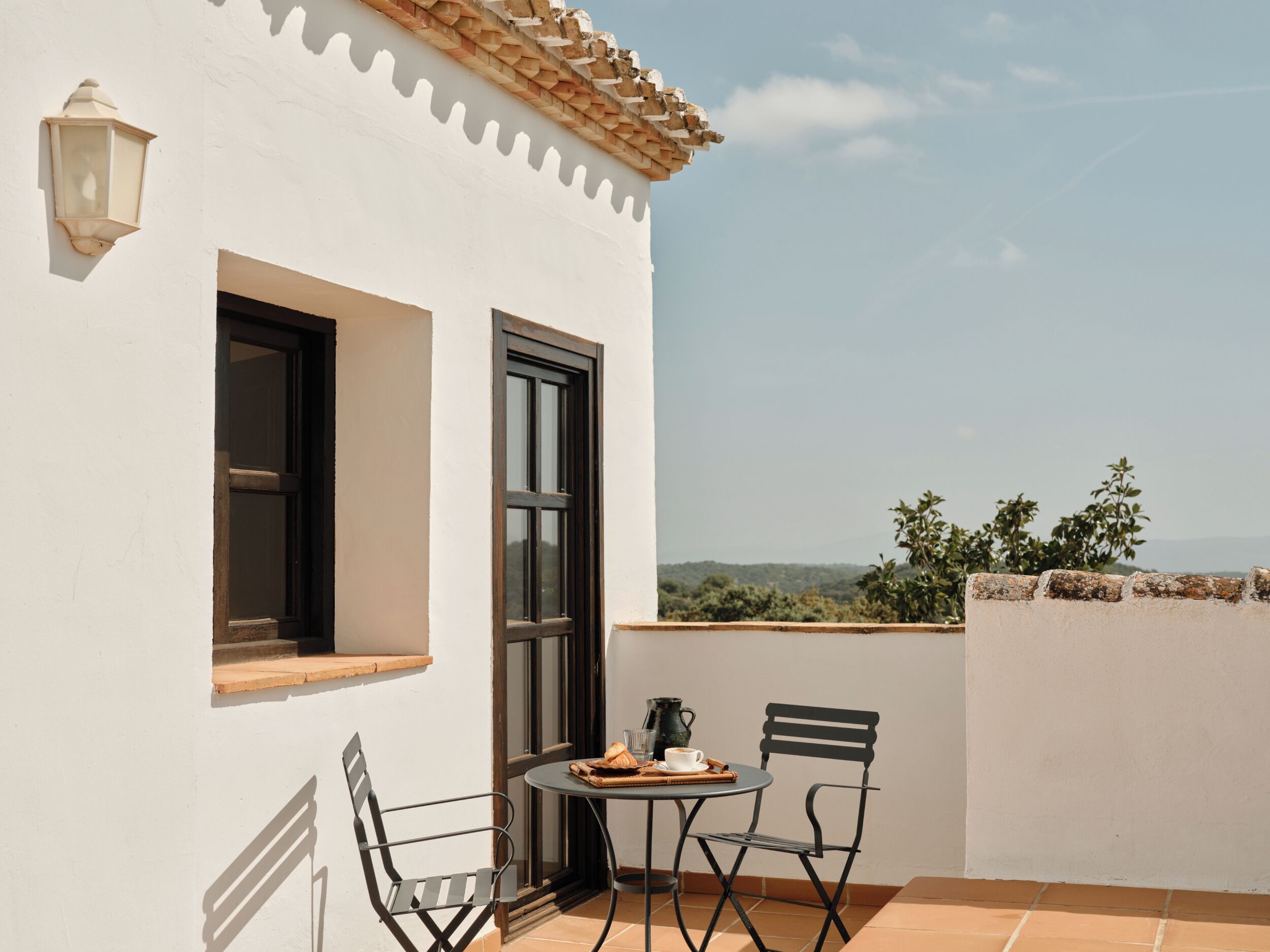 Un bâtiment blanc avec un toit en tuiles, une fenêtre et une porte. On y trouve une table avec des chaises et une petite lampe de table.