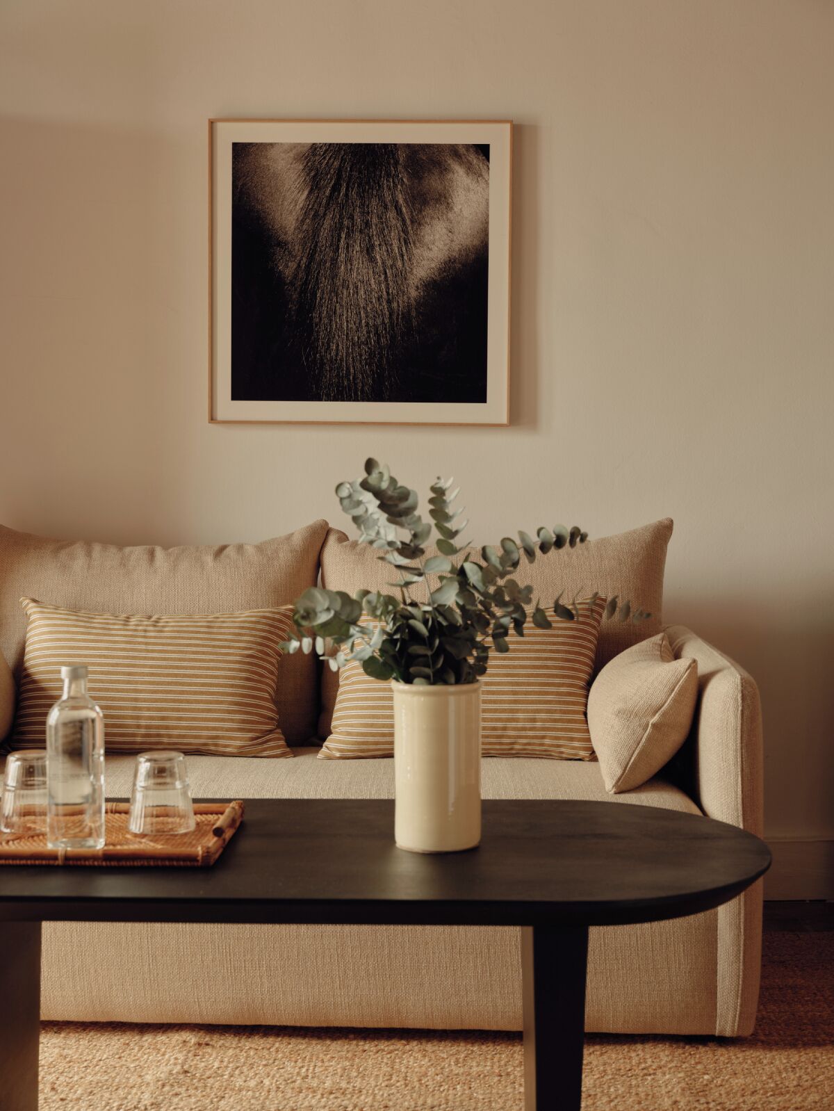 En beige sofa med stribede puder, en vase med grønt løv og et indrammet billede over.
