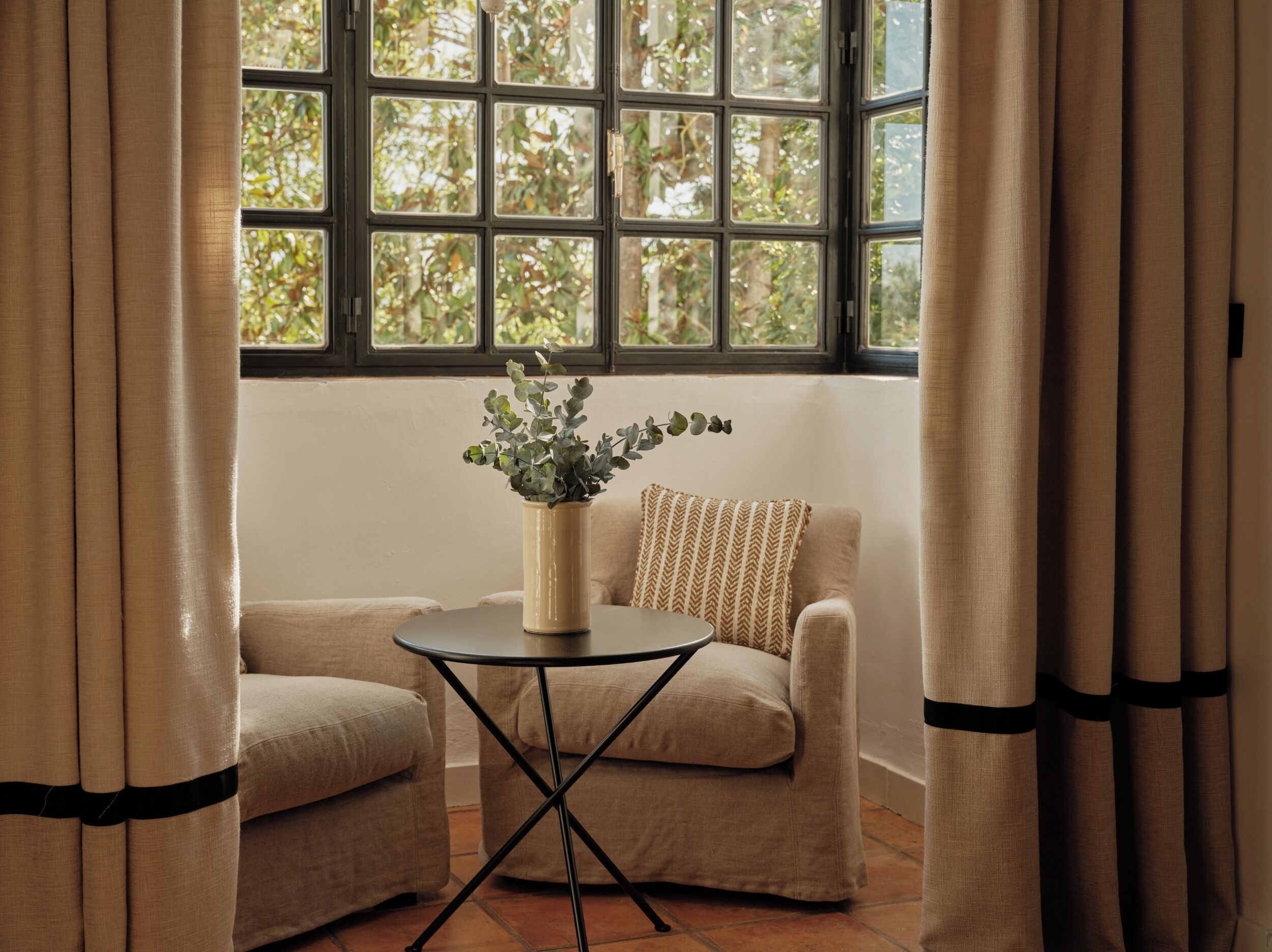 Una habitación con ventana, una pequeña mesa con un florero y dos sillones.