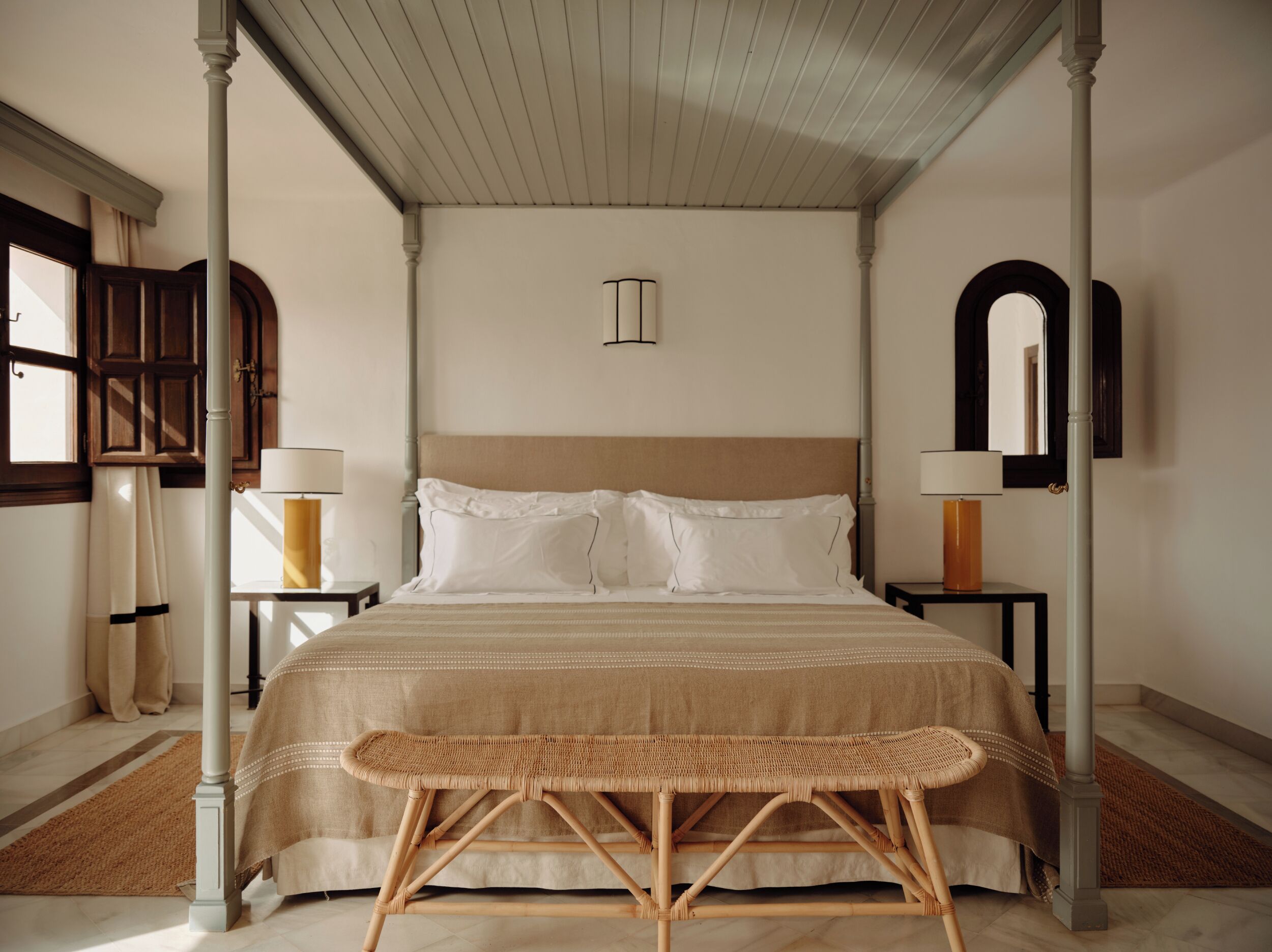 Una cama con dosel con un cabecero color beige, dos mesitas de noche con lámparas y un banco de ratán en una habitación de hotel.