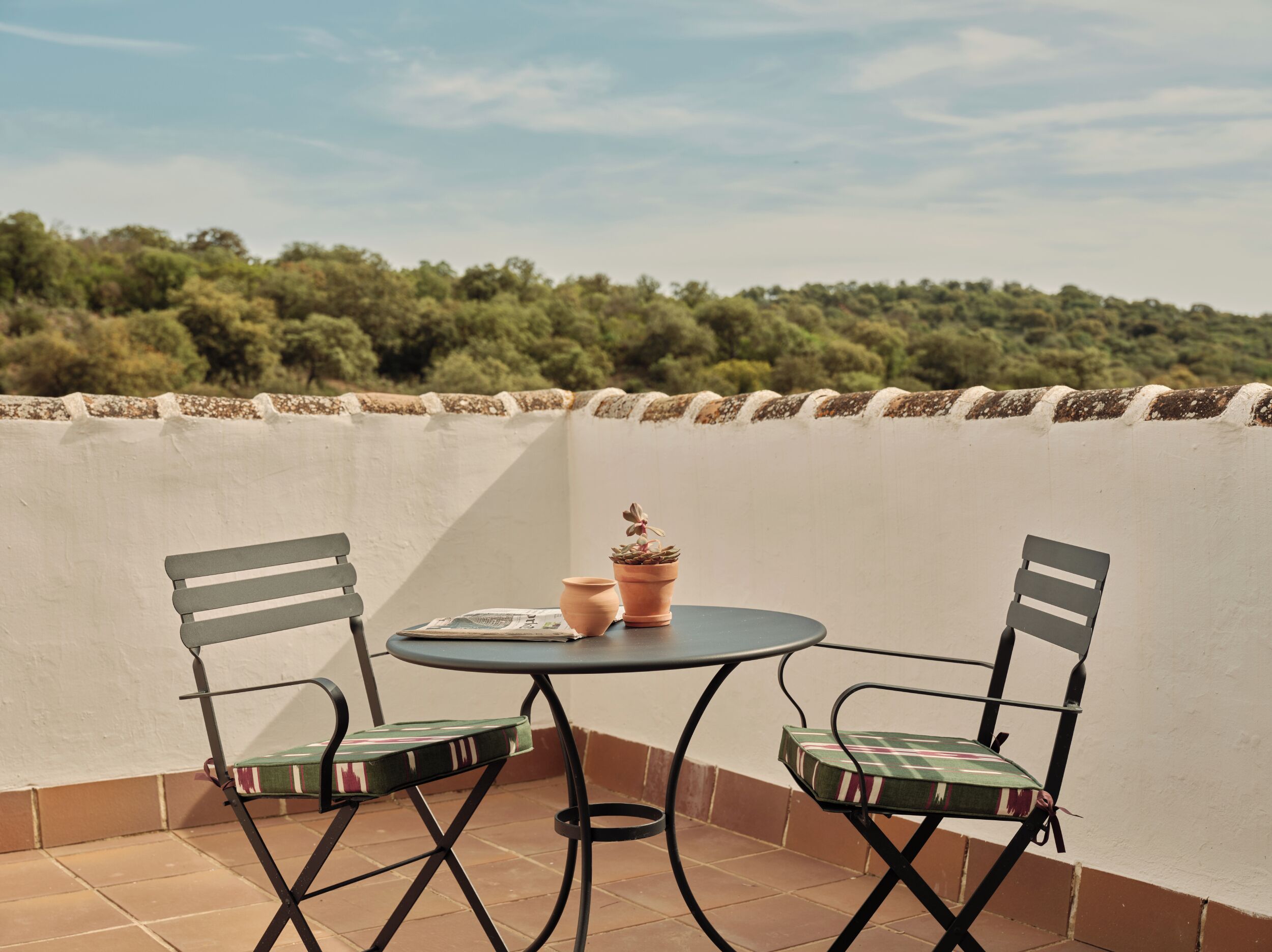 A small outdoor patio with two chairs and a table, surrounded by a white wall and a view of a forested area.