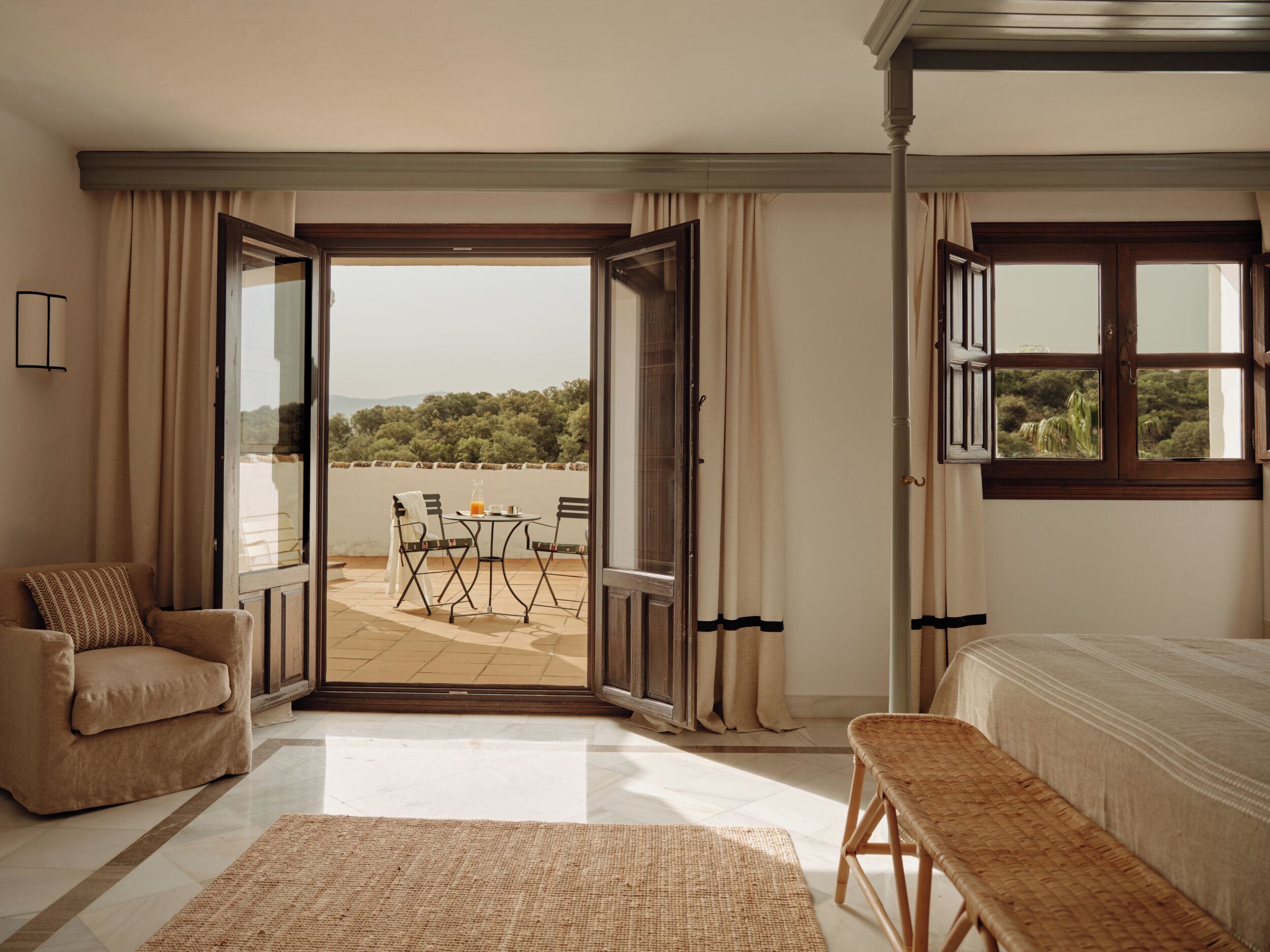 Un dormitorio con cama, un banco, una silla, una mesa con un vaso, y vistas a una terraza con mesa y sillas.